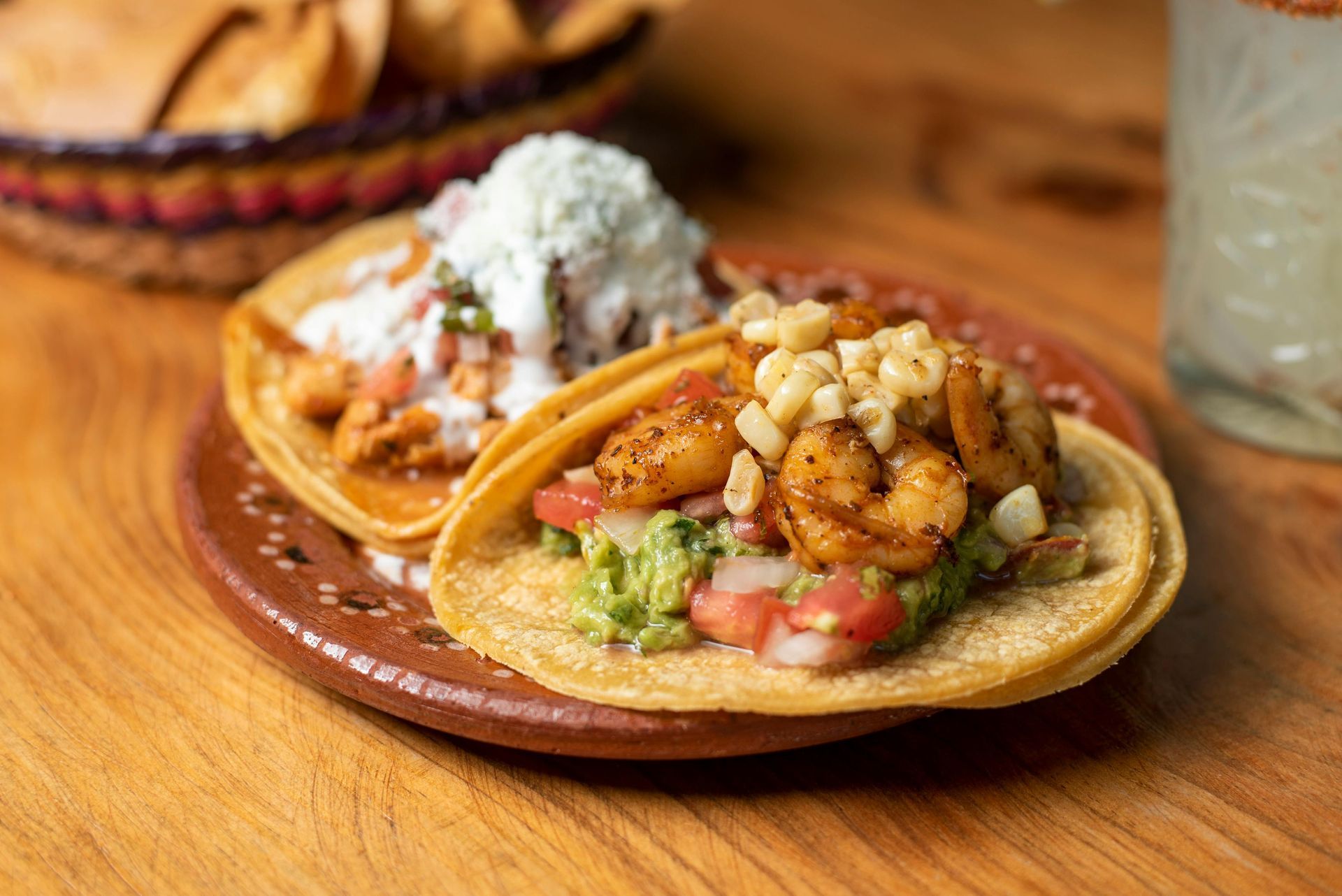 Two tacos on a plate, one with shrimp and corn, the other with chicken and a white topping, with a drink and chips nearby.