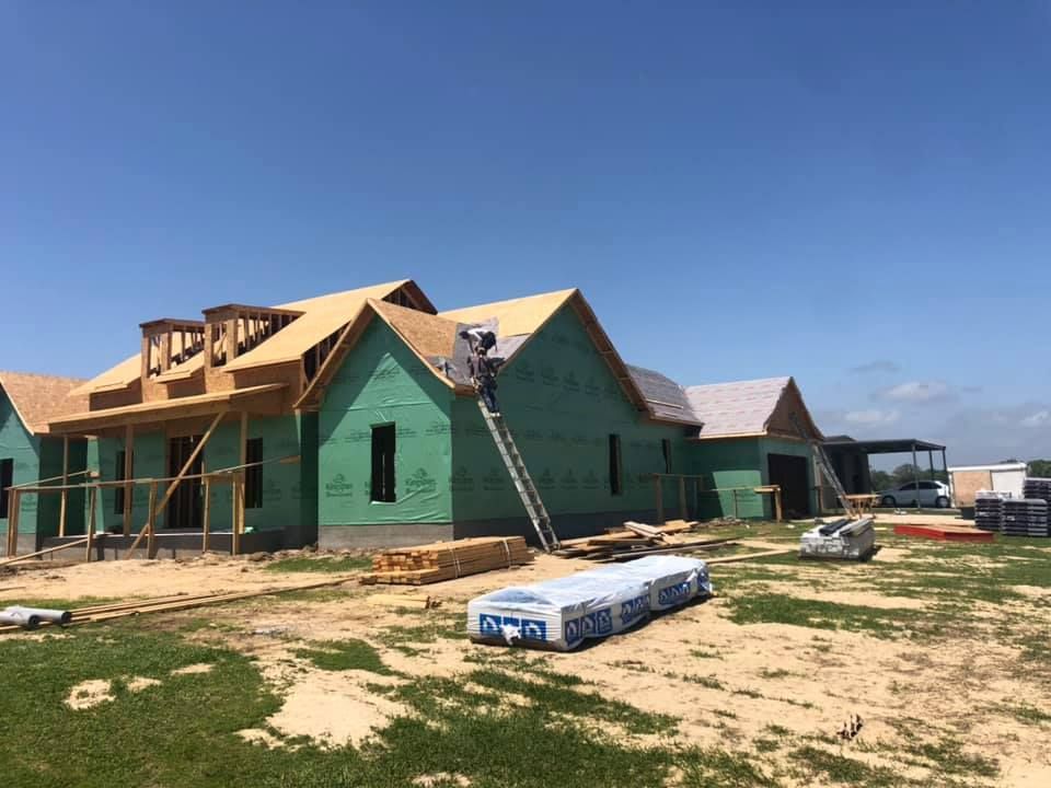 Construction of a house with green exterior, wood beams, and a worker on a ladder, under a blue sky.