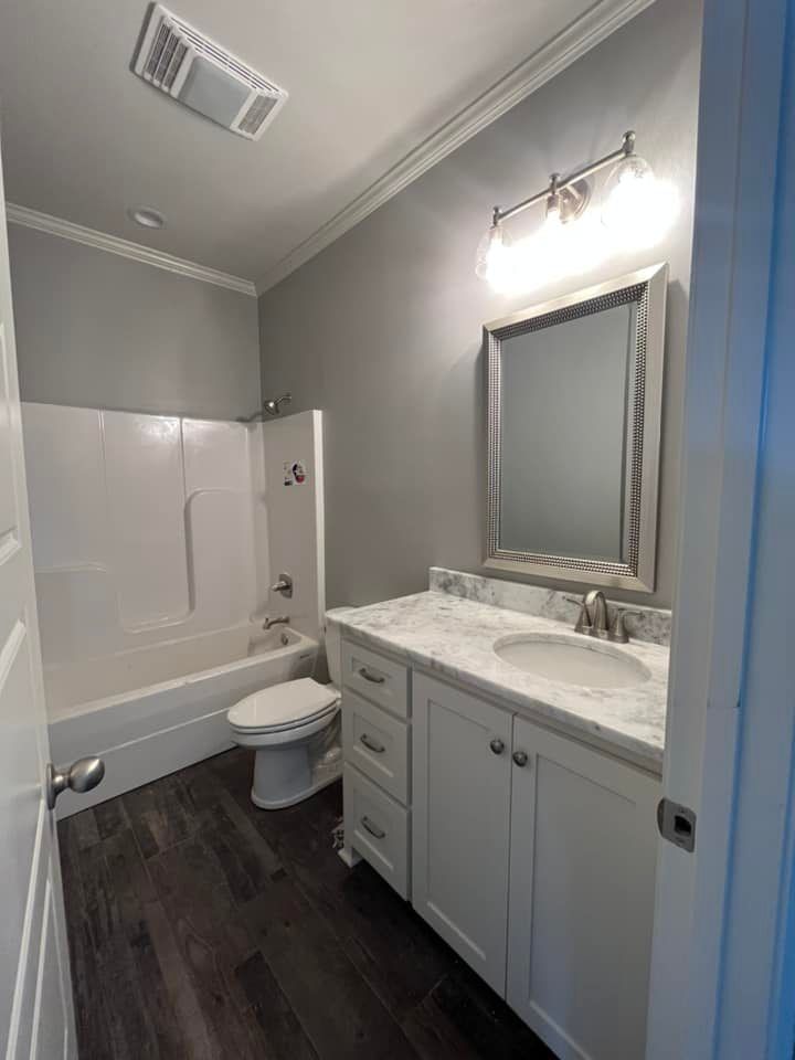 Bathroom with white vanity, tub, and toilet. Gray walls, dark wood-look floor, and silver-framed mirror.