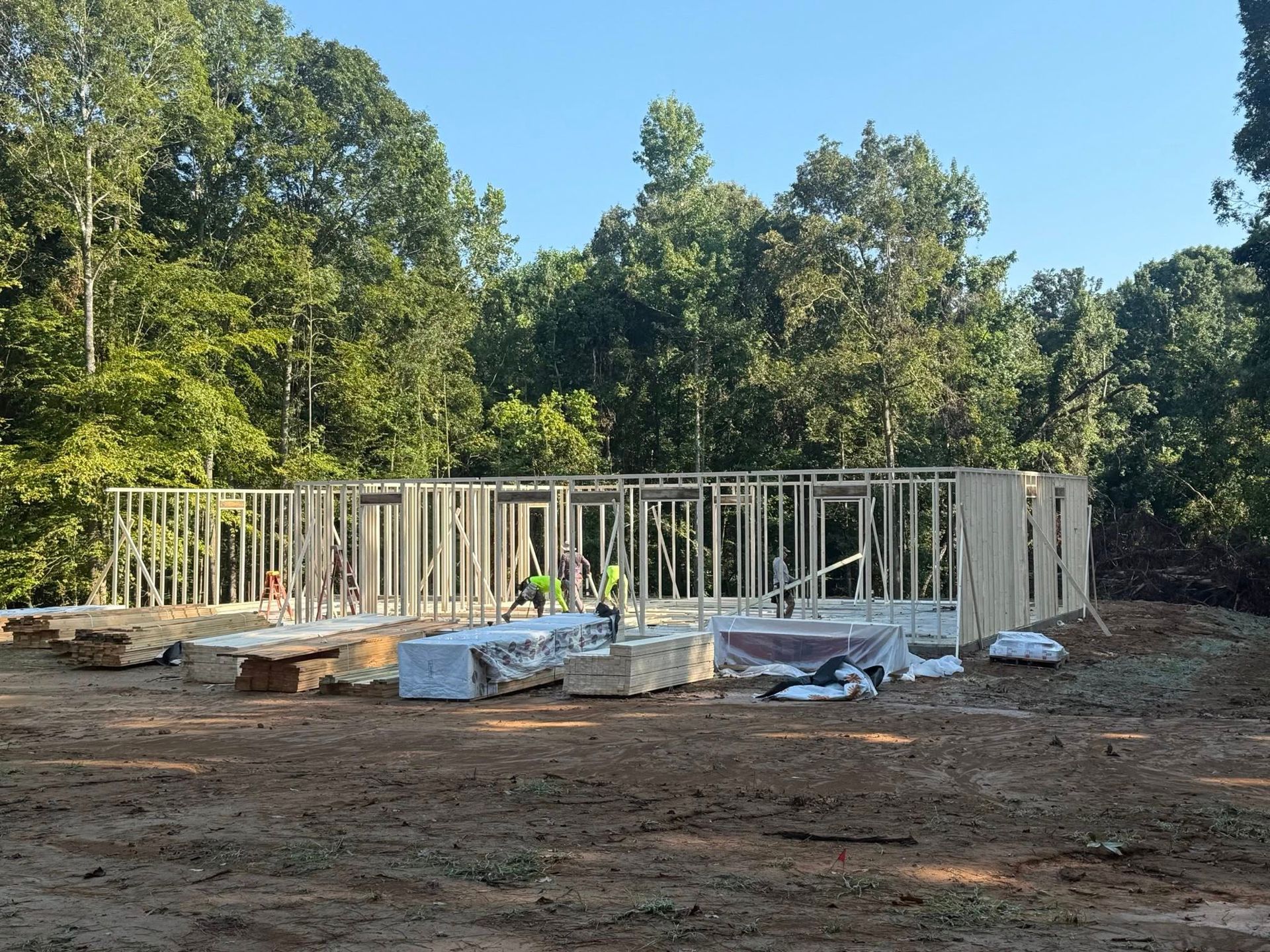 Framing of a house under construction in a wooded area with workers present.