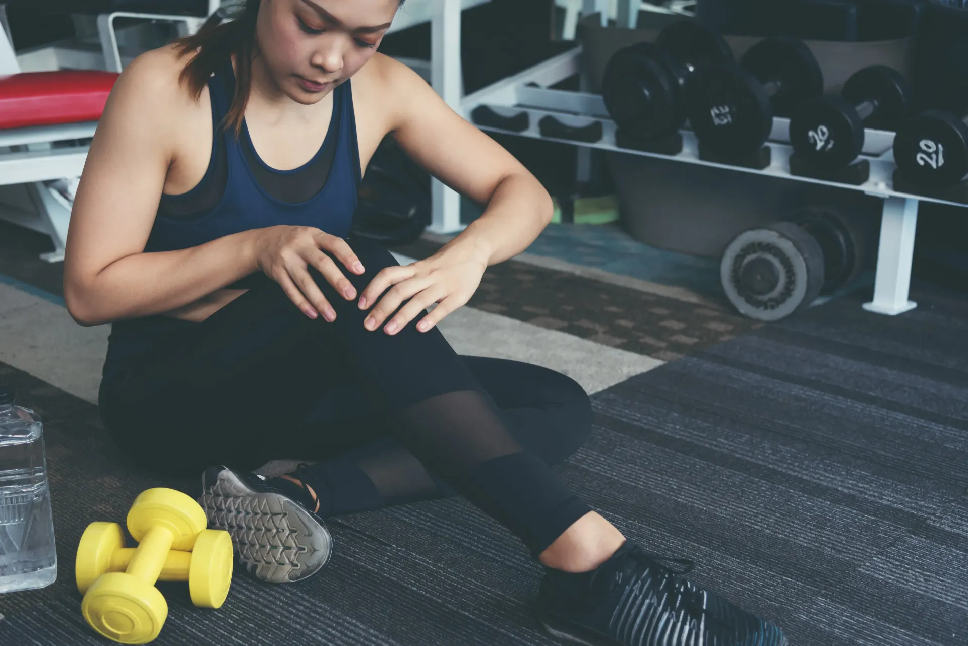 Woman in workout clothes at gym, holding knee, possibly injured. Yellow dumbbells on floor.