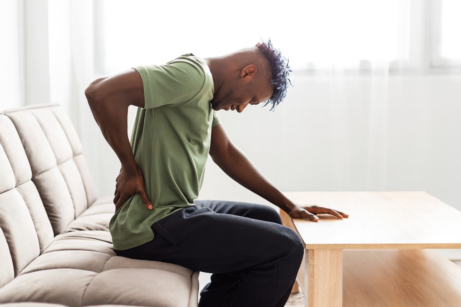 Man seated, grimacing, clutching his lower back in pain, indoors near a window.