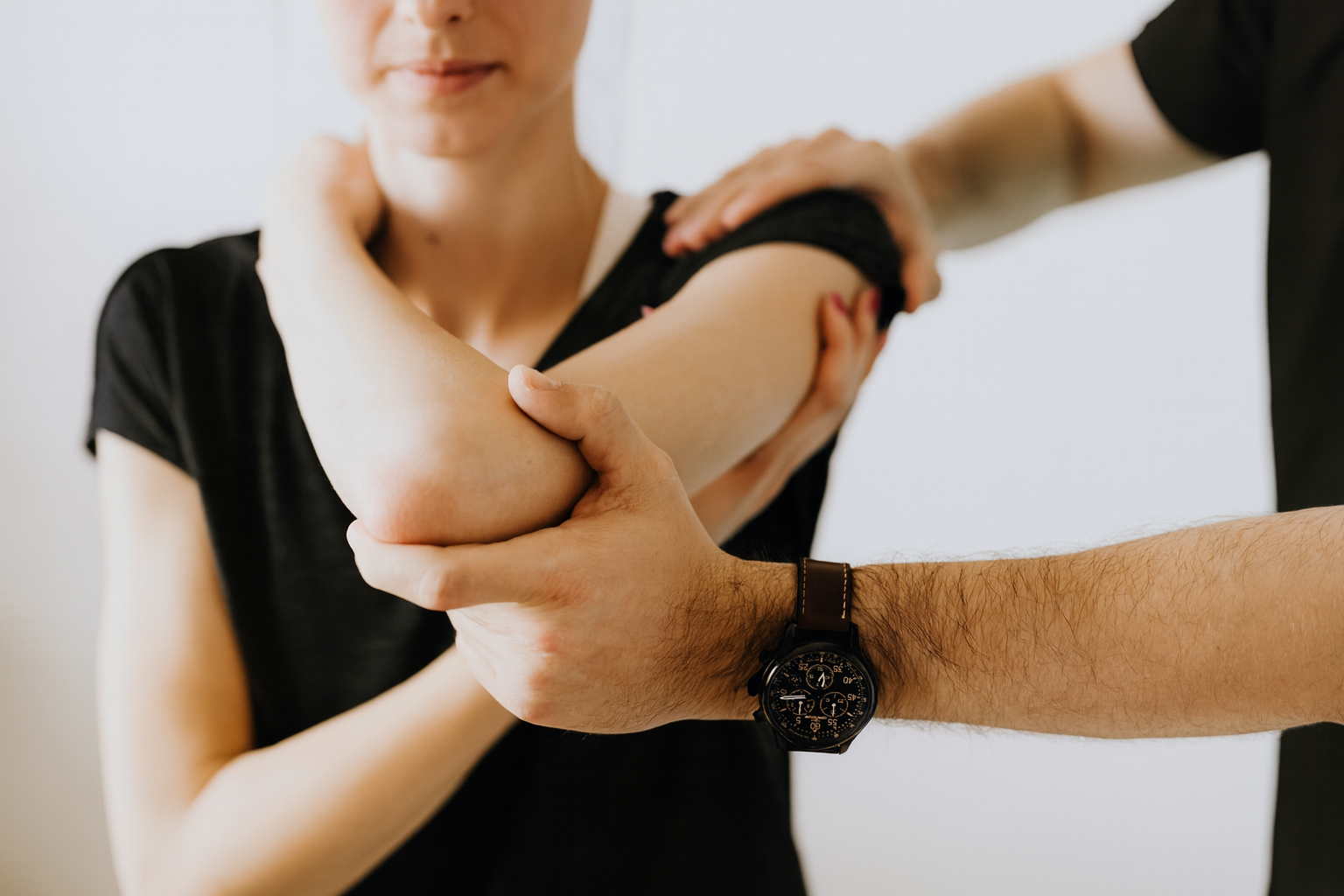 A person's elbow being examined by another person. Both are wearing black shirts; the setting is a neutral space.