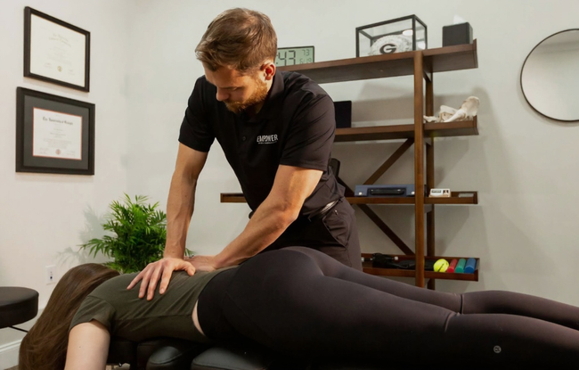 Chiropractor adjusting patient's back in a clinic; woman laying prone, man wearing black shirt applying pressure.