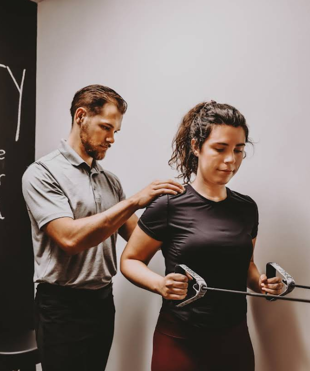 A man assists a woman with an exercise using resistance bands; indoors, white wall.