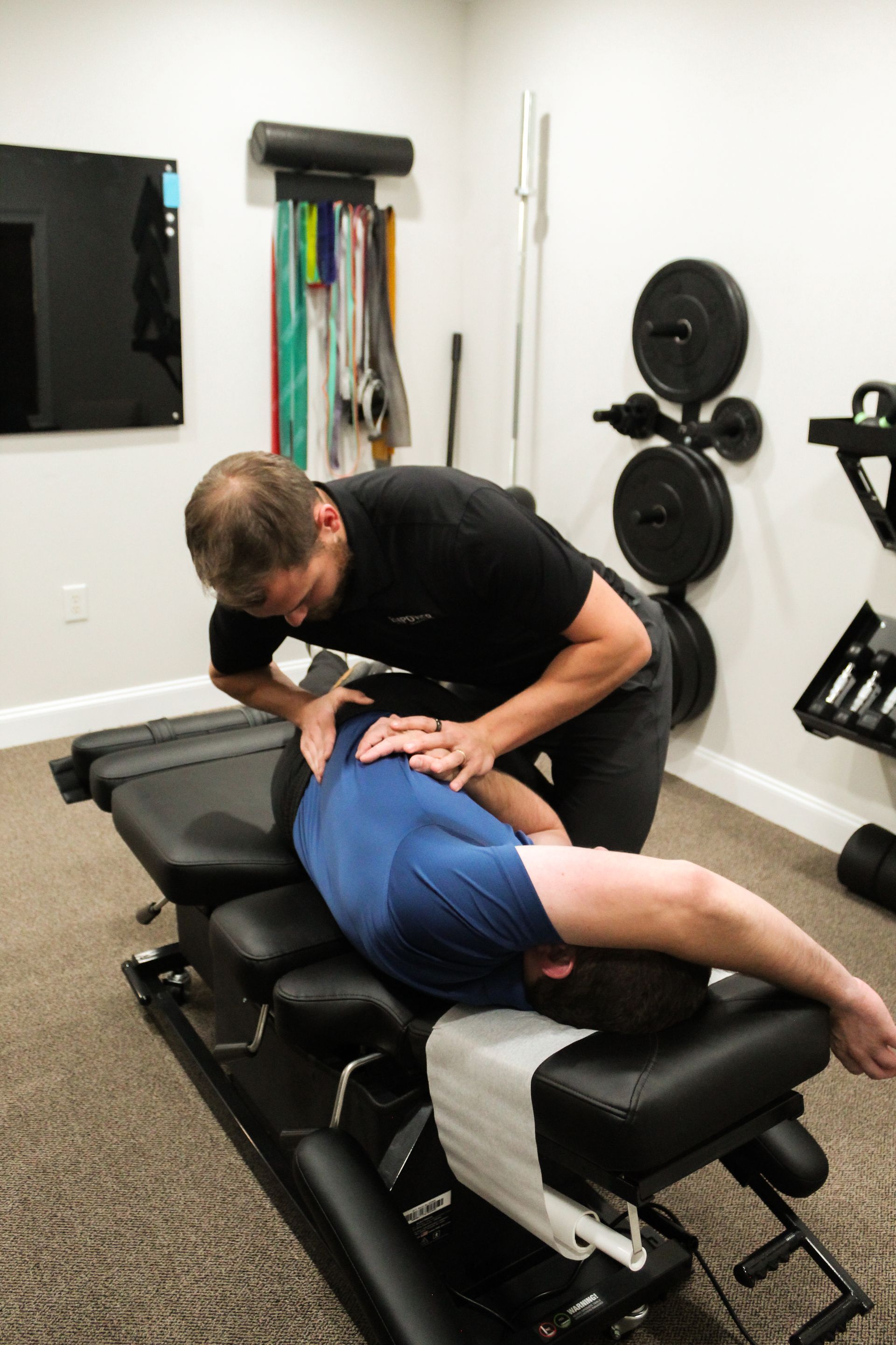 Man lying face down on a massage table, wearing yellow shorts and a black shirt, in a clinical setting.
