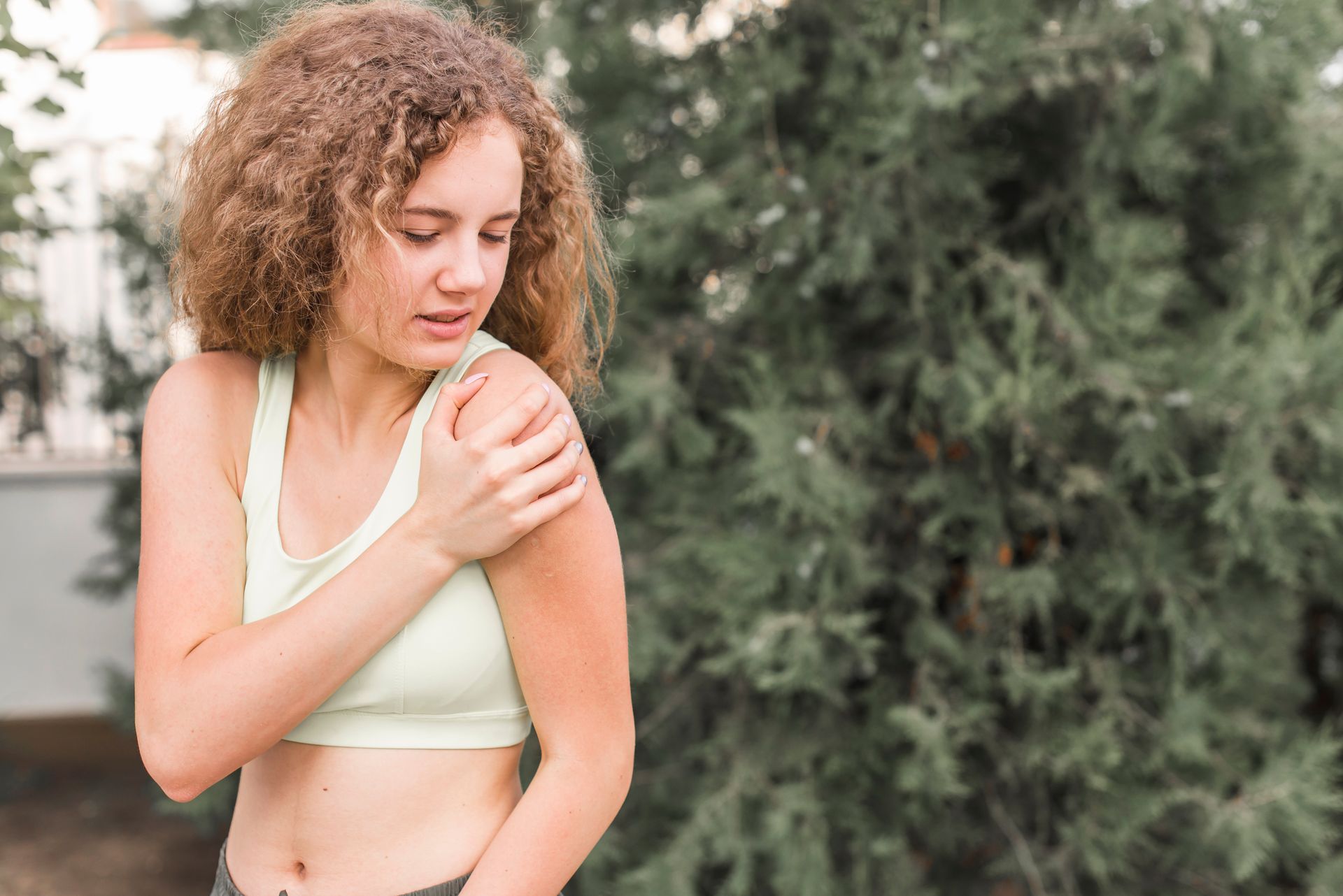 Woman with curly hair holding her shoulder, looking down in pain, outside.