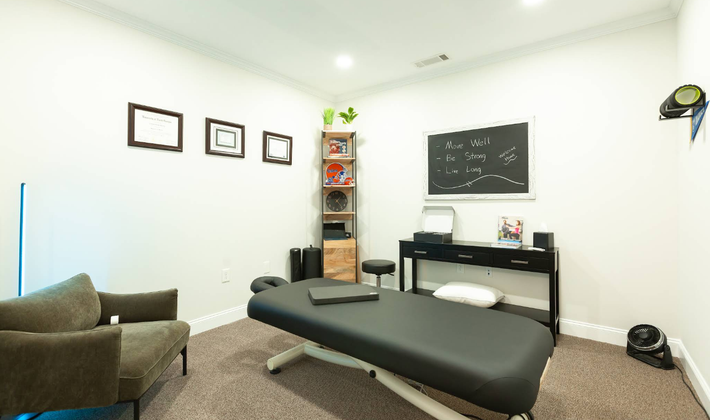 A therapist's office with a massage table, armchair, desk, and framed certificates on the wall.