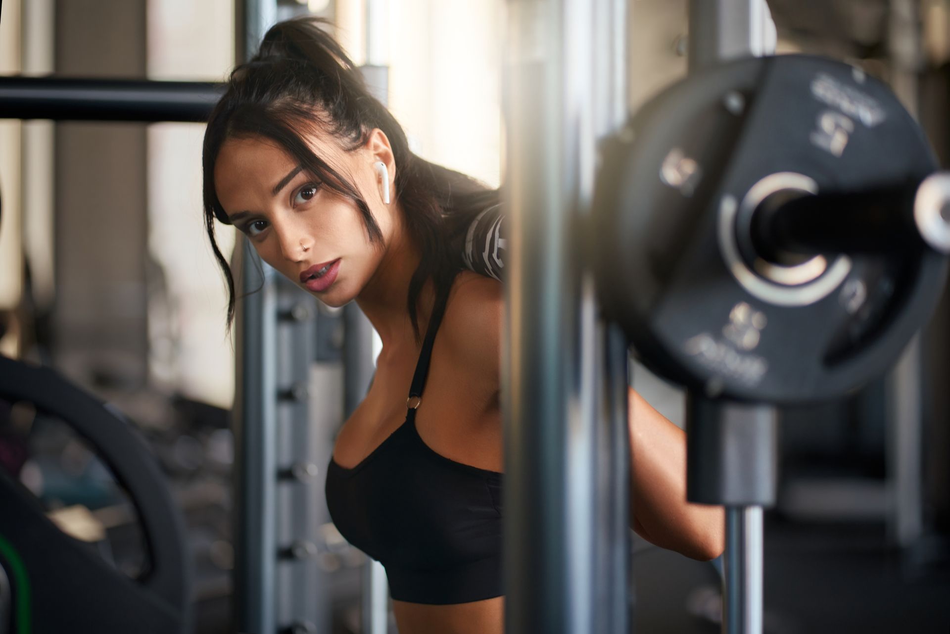 Woman in black sports bra at the gym, looking over her shoulder.  Barbell in the foreground.