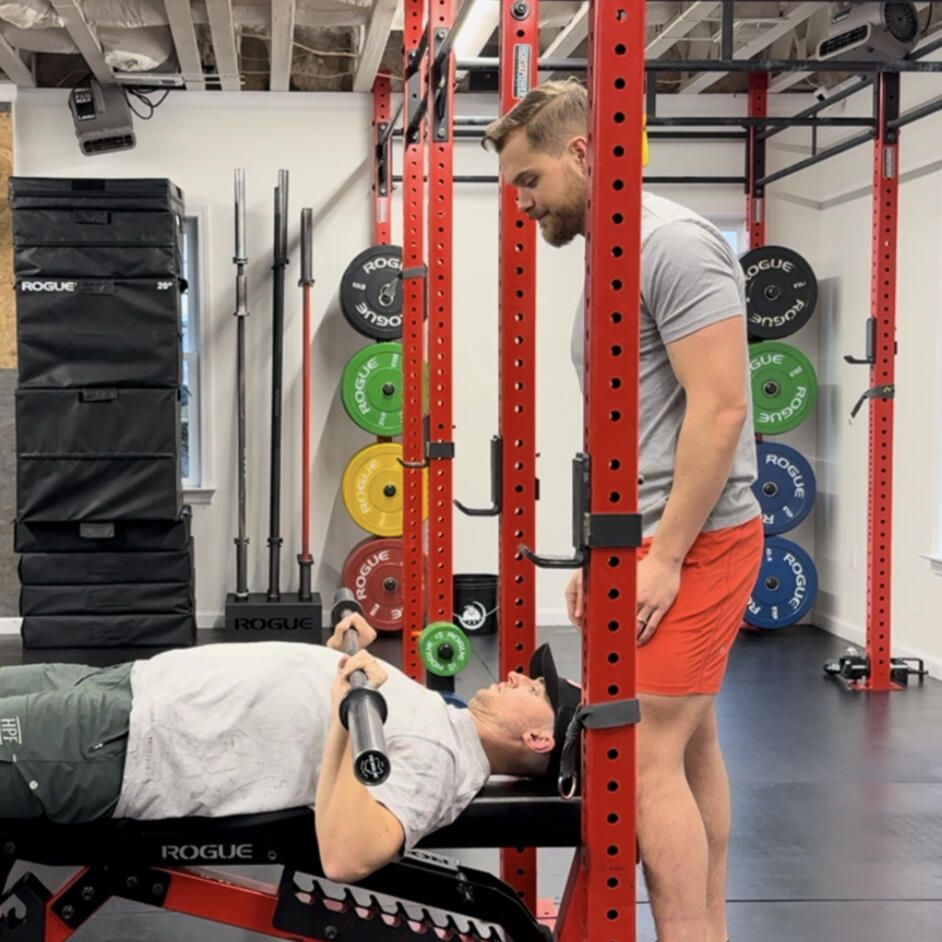 Man on weight bench, lifting bar with spotter in a gym. Red weight rack, weights, and fitness equipment are present.