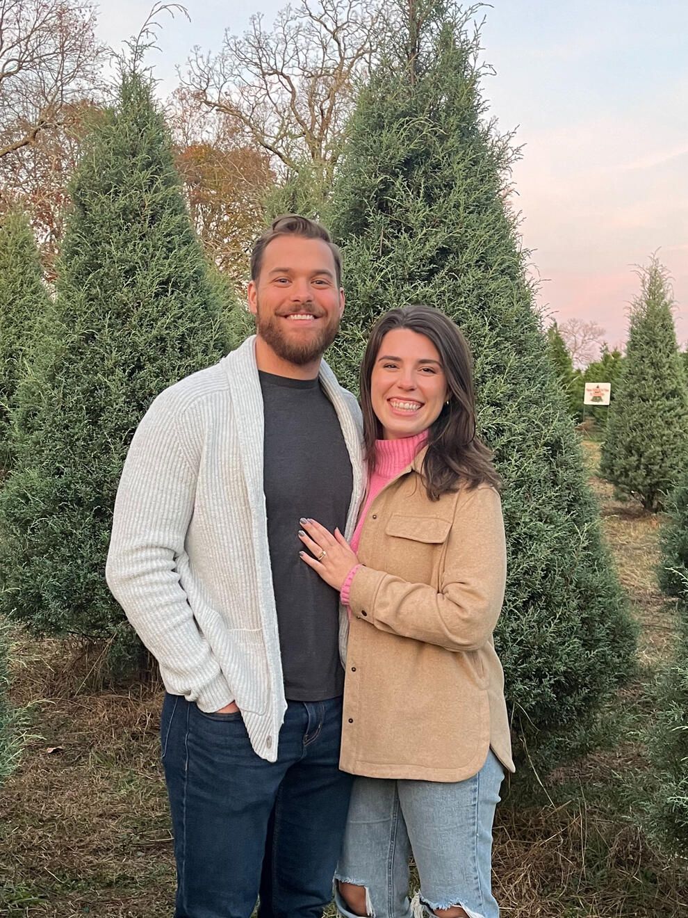 Couple poses outdoors in front of evergreen trees. Man wears a white sweater, woman in a tan jacket and pink shirt.