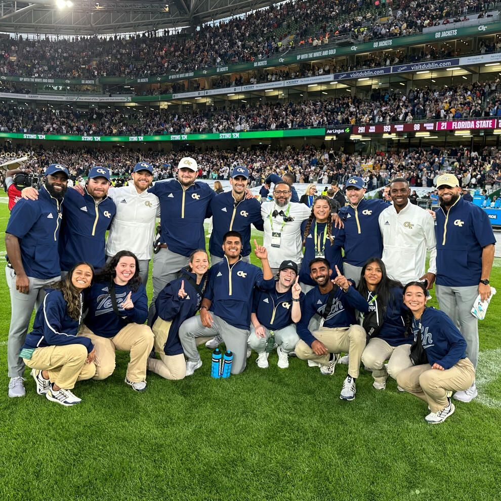 Group of people on a football field, in navy and gold, smiling, celebrating a game. Large stadium crowd in background.