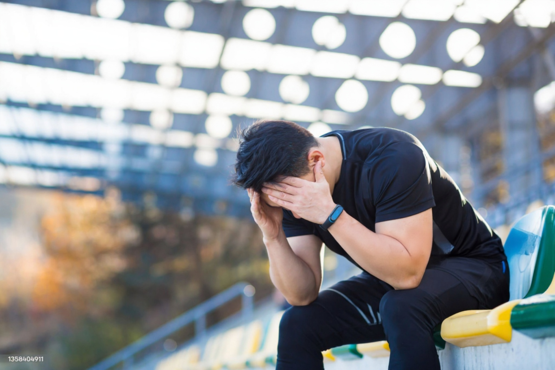 Man in black sportswear with head in hands, sitting on bleachers in a stadium, looking distressed.