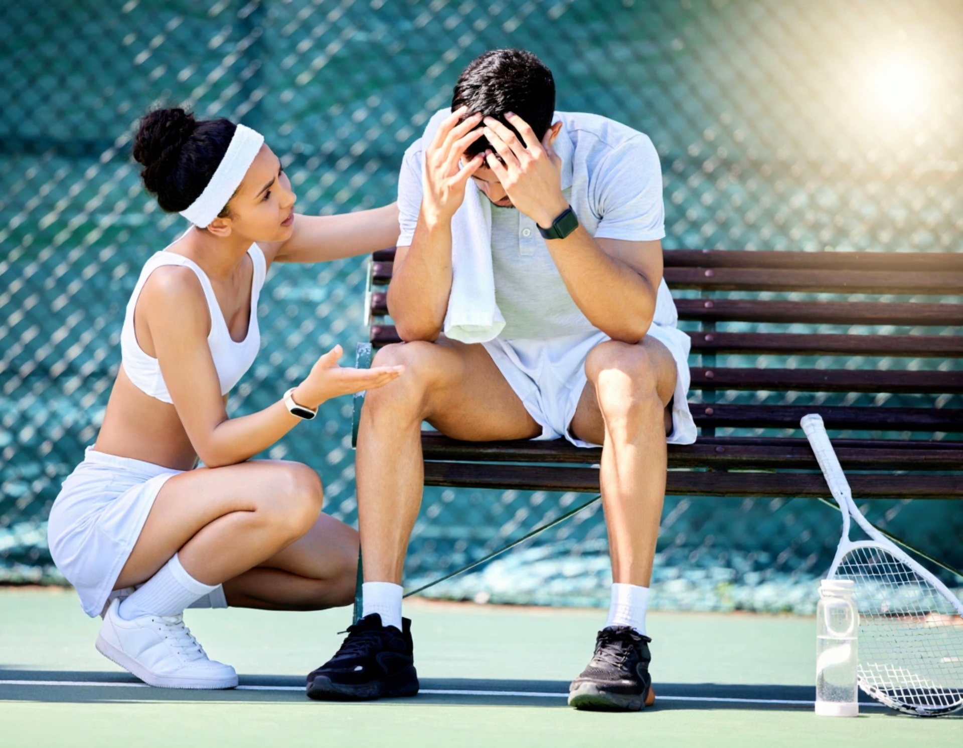 Woman comforts a distressed man on a tennis court bench. He holds his head, towel in hand.