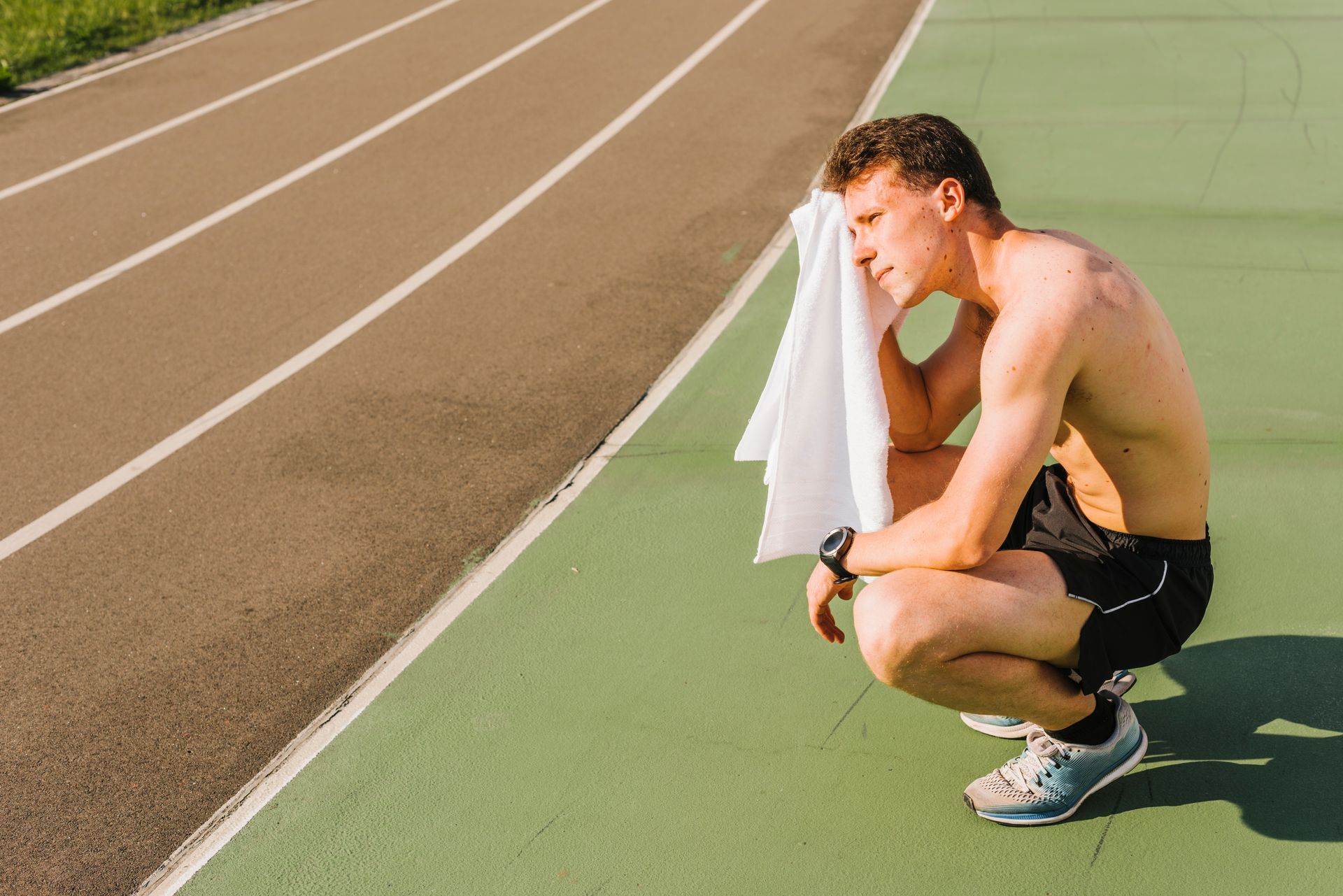 Man in athletic wear resting on a track, wiping sweat with a towel.