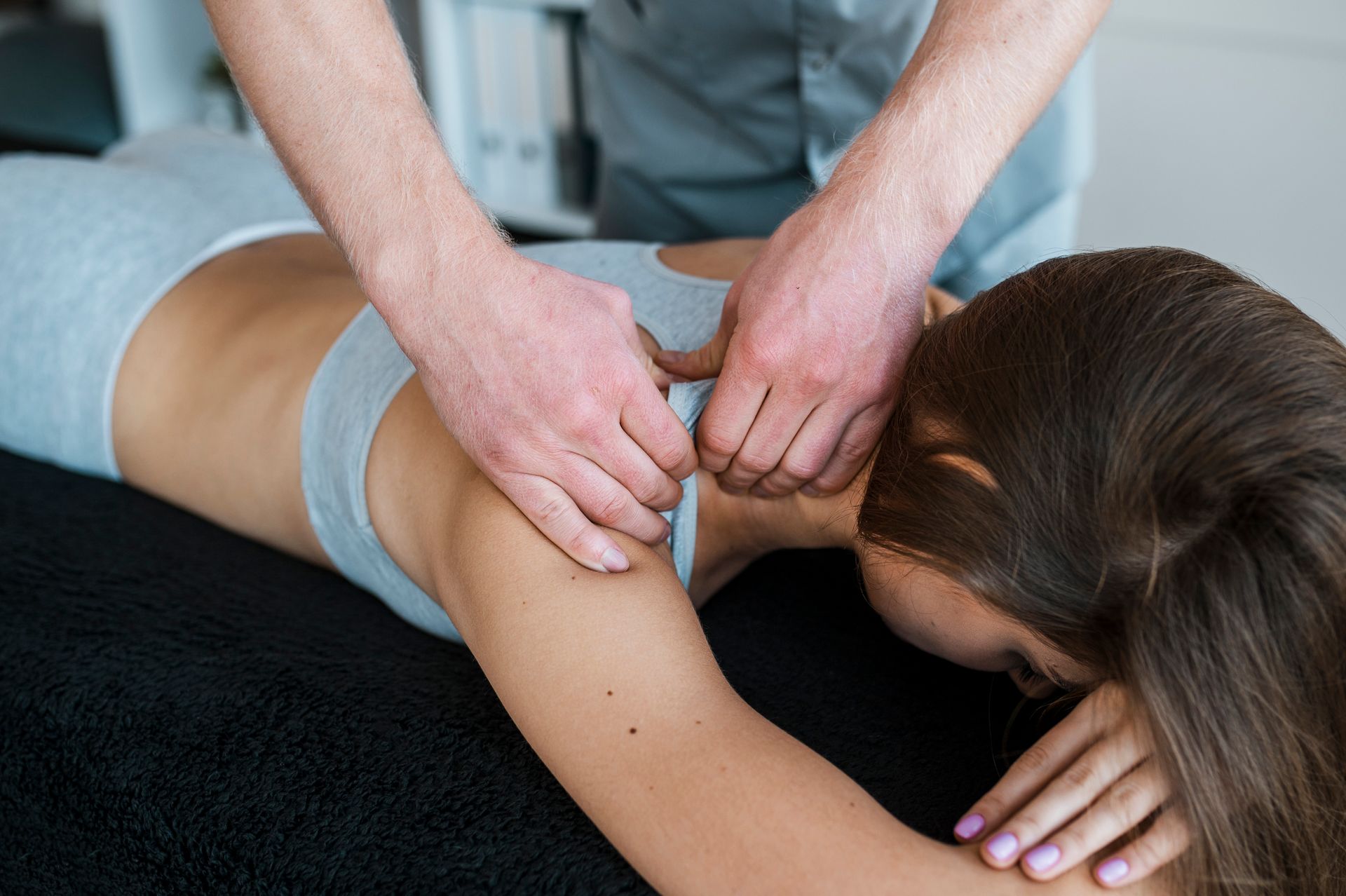 Person receiving shoulder massage on a black surface. A therapist's hands knead the person's shoulder. Both are wearing gray.
