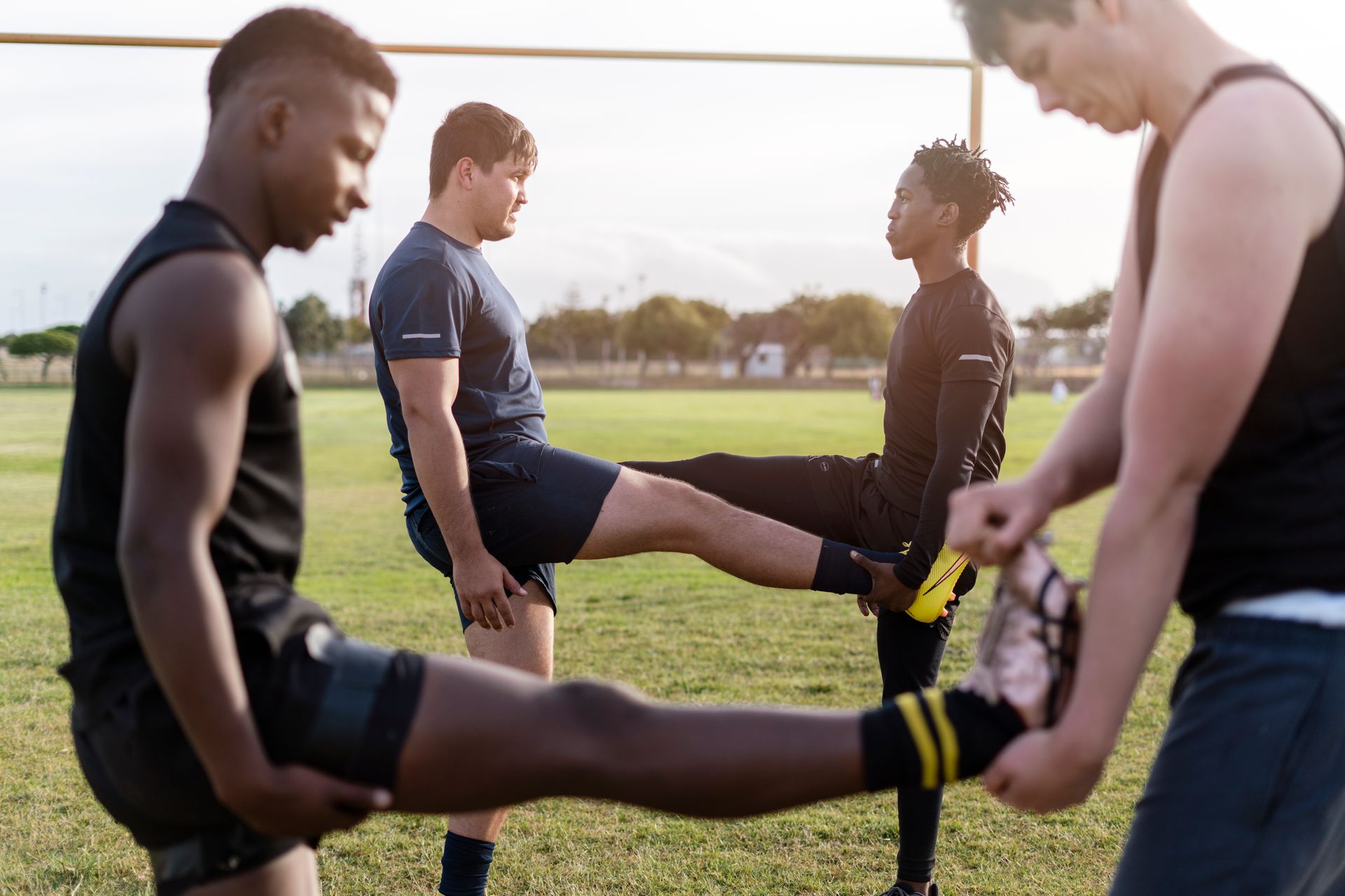 Four athletes stretching legs on a green field, one holding the other's foot.