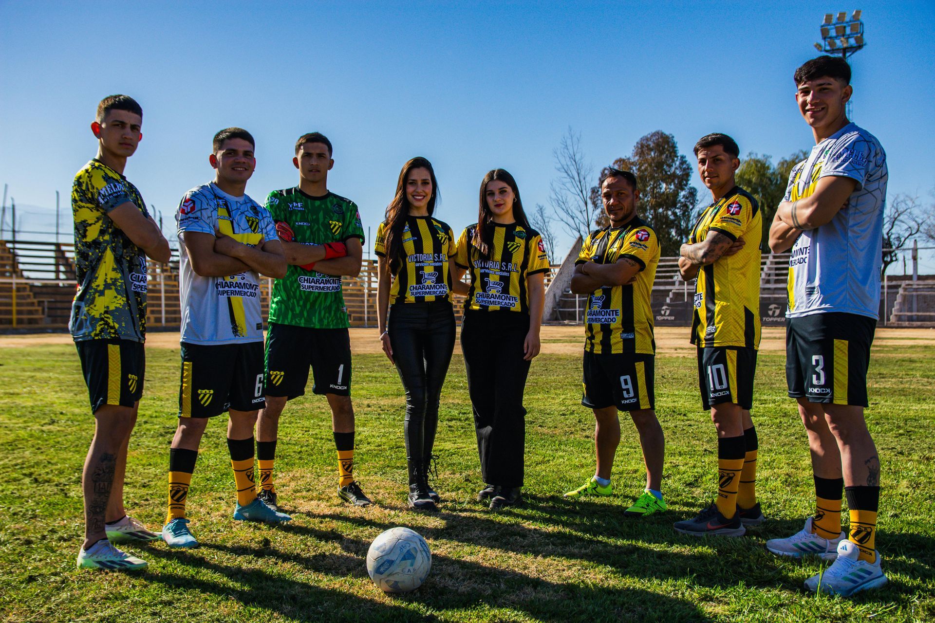 Soccer team poses on a field. Players in black, yellow, and green jerseys. A soccer ball is in front.