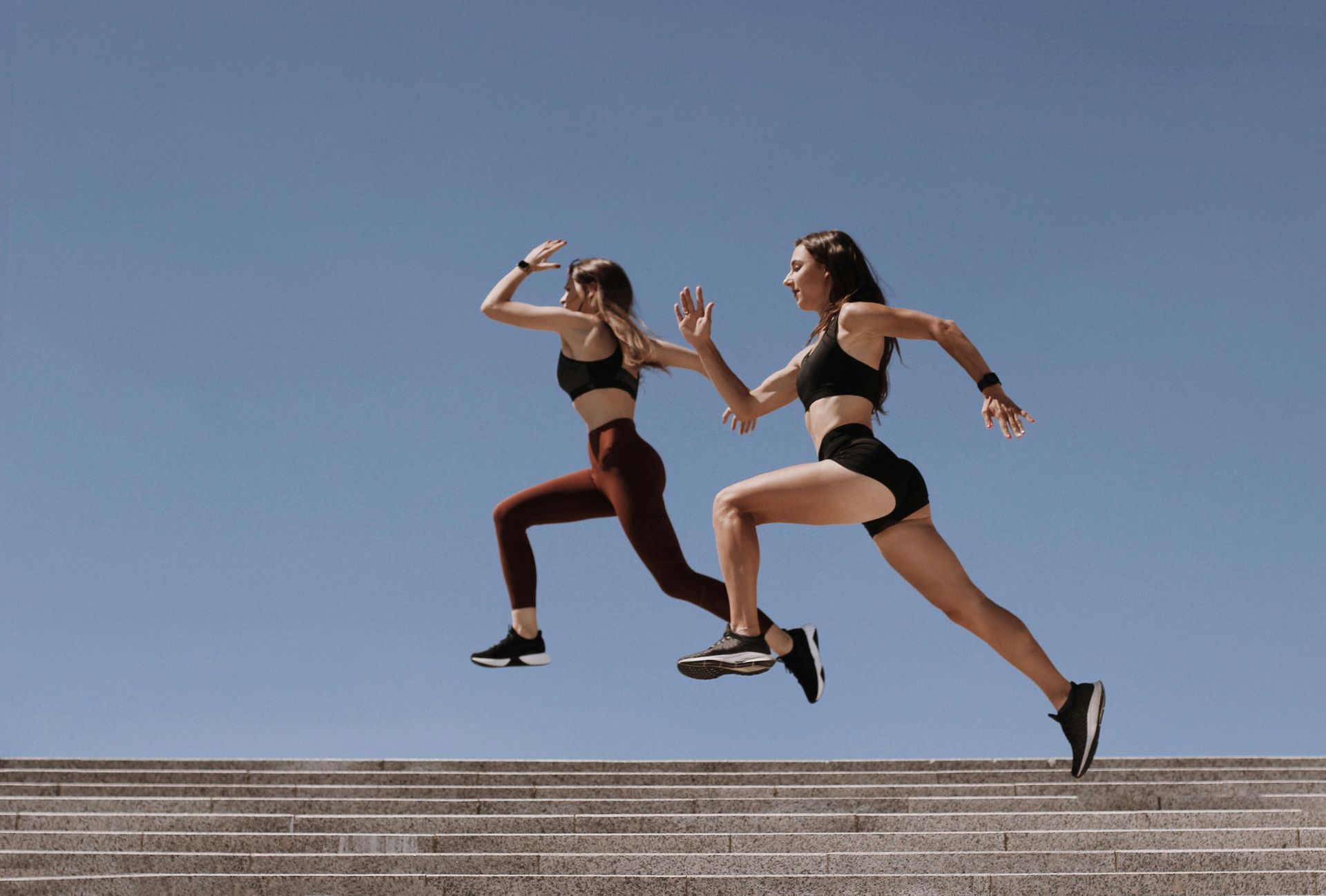 Two women running up stairs under a blue sky. One wears brown leggings, the other black shorts and sports bras.