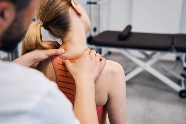 A healthcare professional examining a woman's neck and back, possibly for pain. Indoors, light-skinned patient and practitioner.