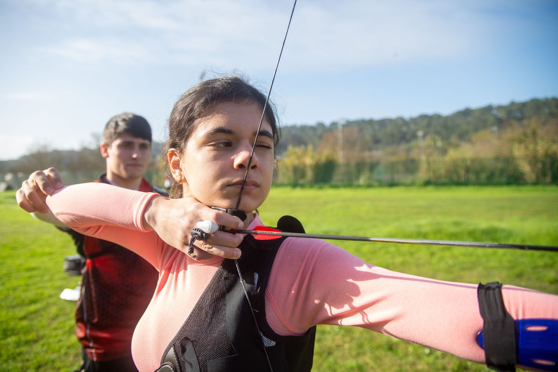 Person drawing a bow and arrow outdoors on a sunny field, with another person in the background