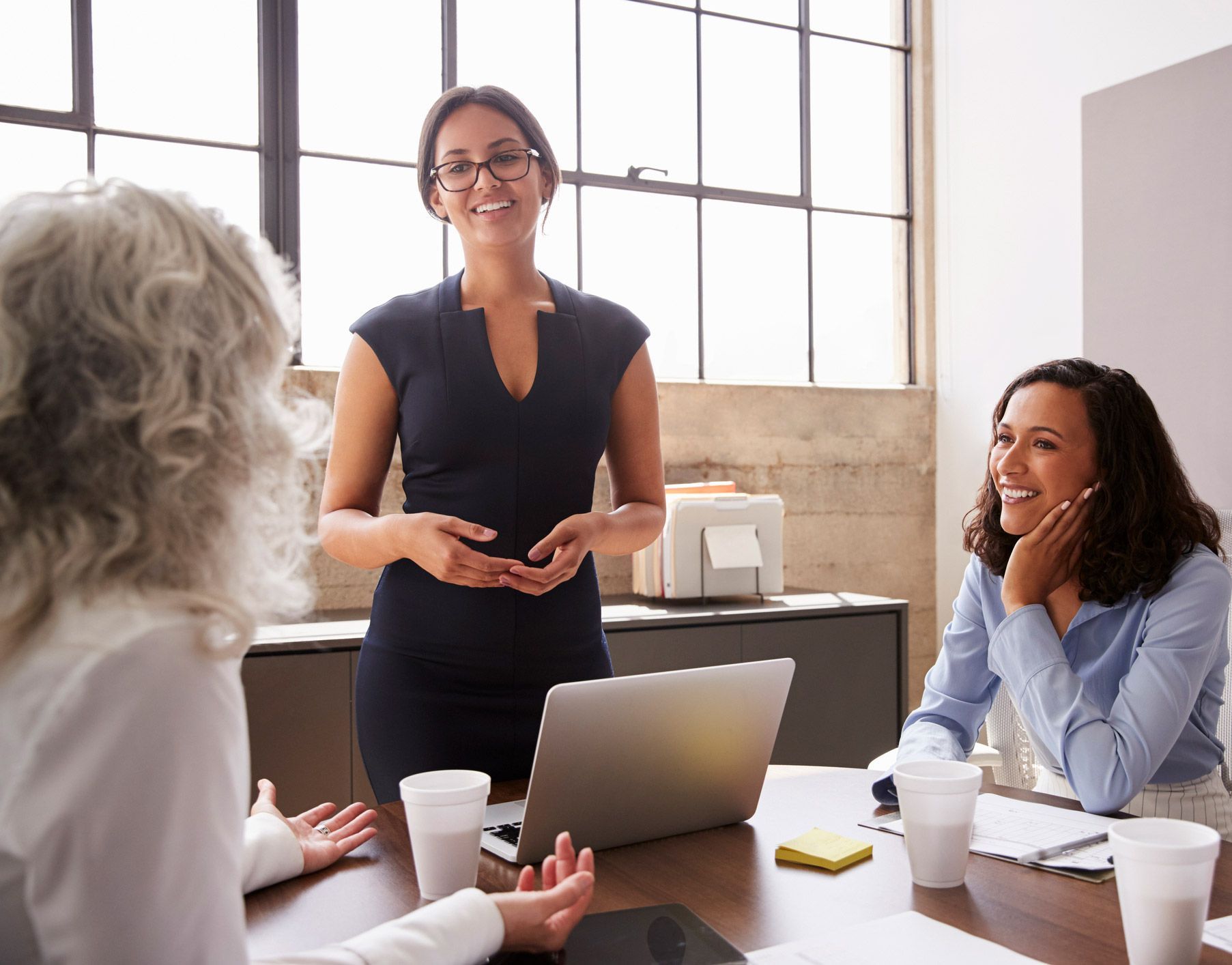 Woman in navy dress presents to colleagues at a meeting table.