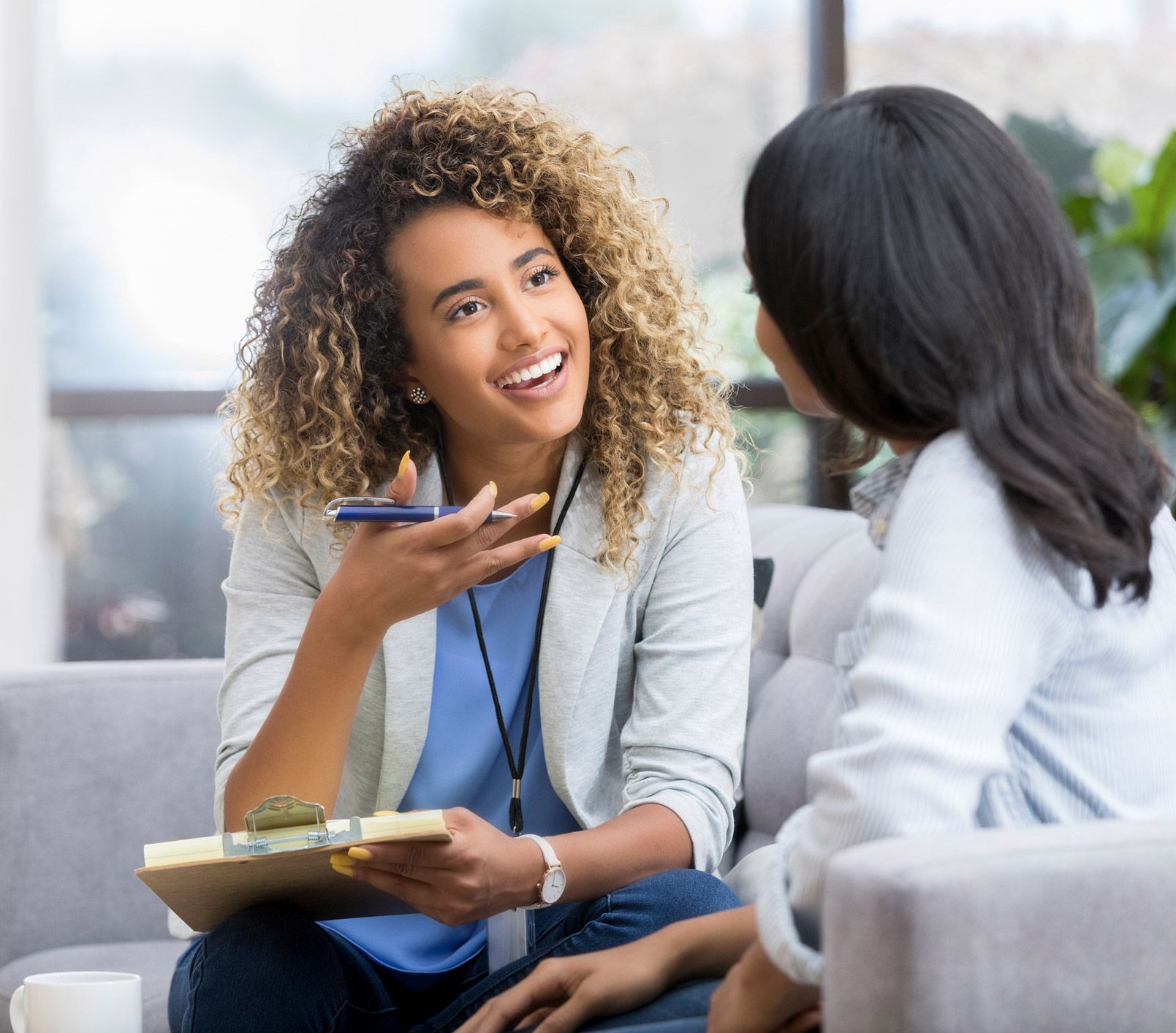 Woman with curly hair talks to another woman, holding a clipboard, indoors.