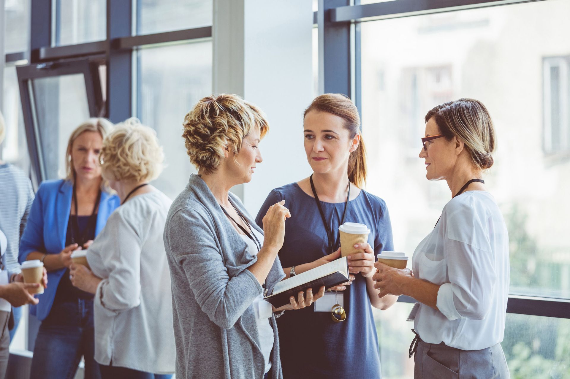 Group of women in office setting, conversing, holding coffee cups. Natural light, large windows.