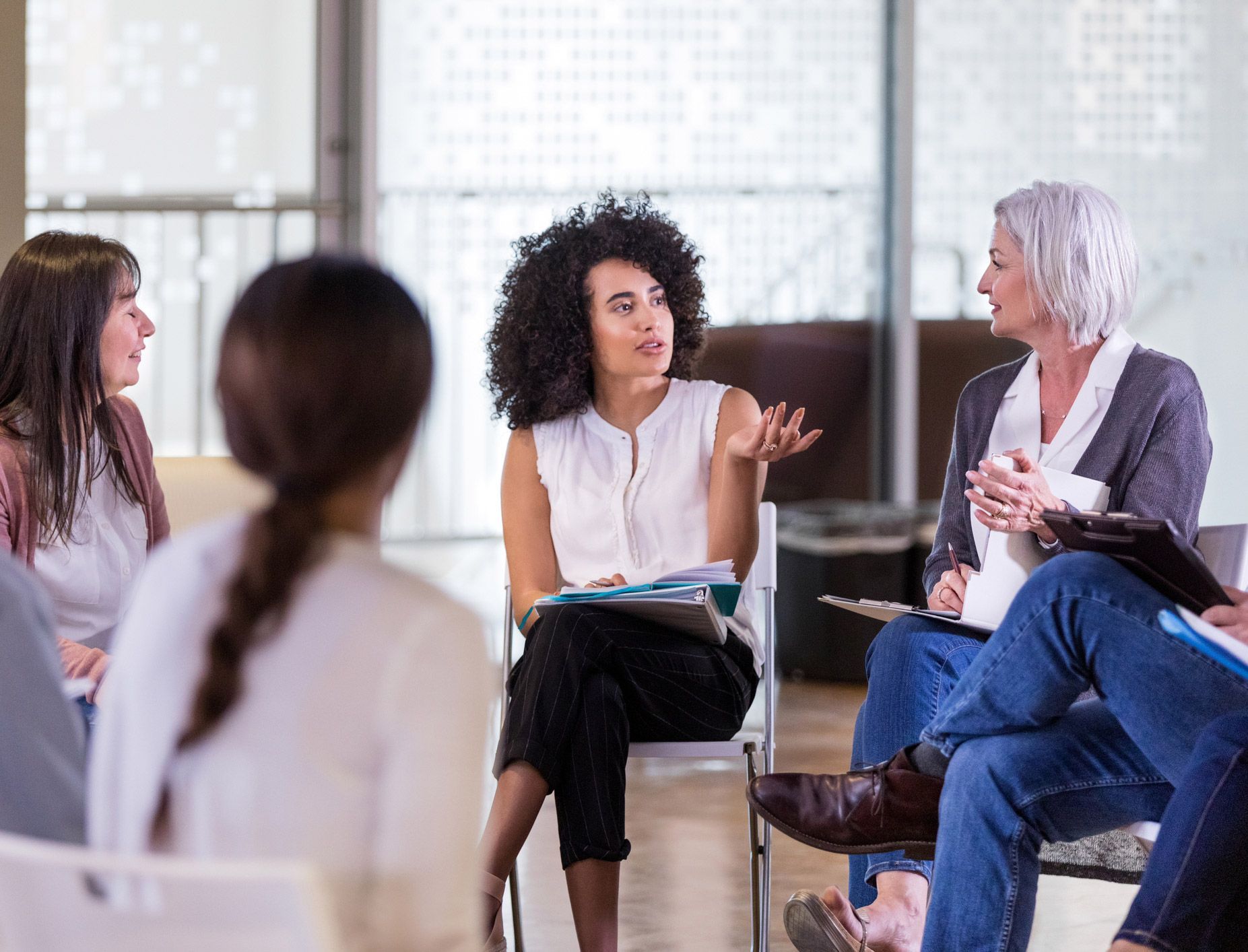 People in a circle, engaged in a discussion; one woman gestures, another listens, holding a notepad.