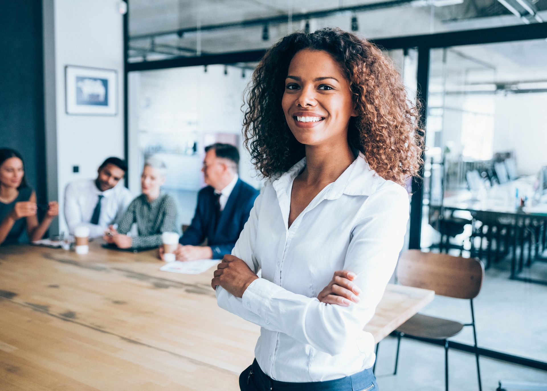 Woman in white shirt, arms crossed, smiling in a conference room with colleagues in the background.