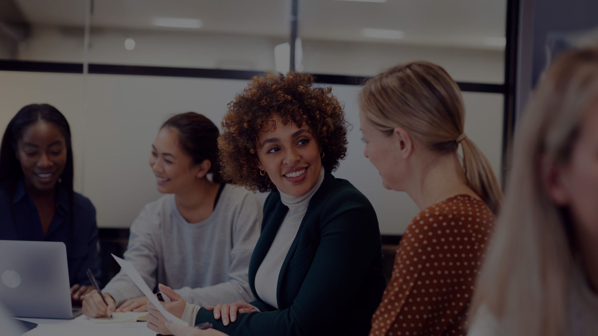 Women at a table in a meeting, smiling and looking at documents. Inside a modern office.