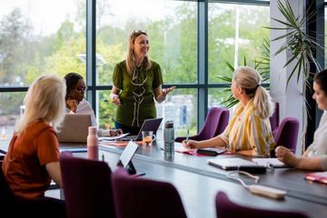 Woman in green top leads meeting at long table with other women in a modern office.