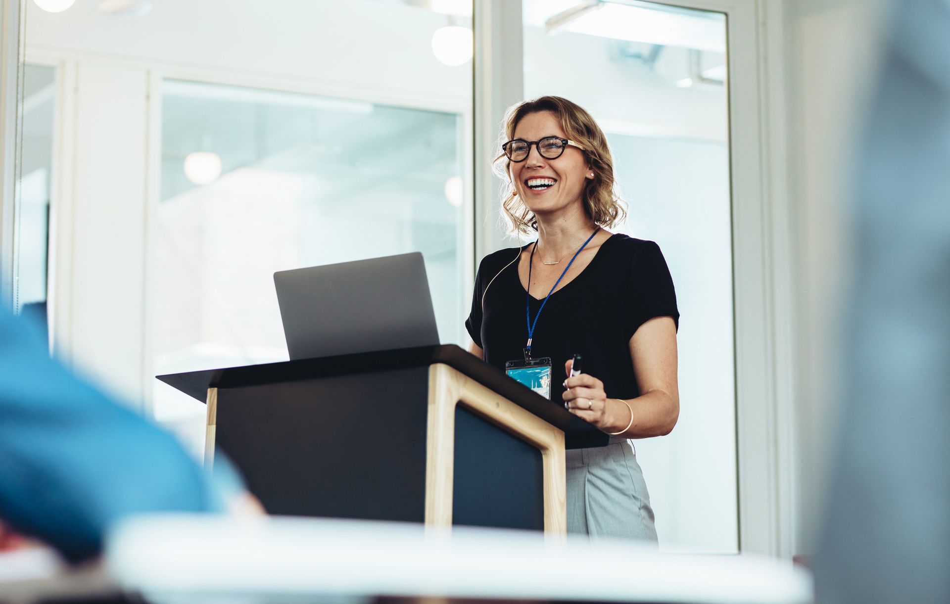 Woman smiling, presenting at a lectern with a laptop in a conference room.