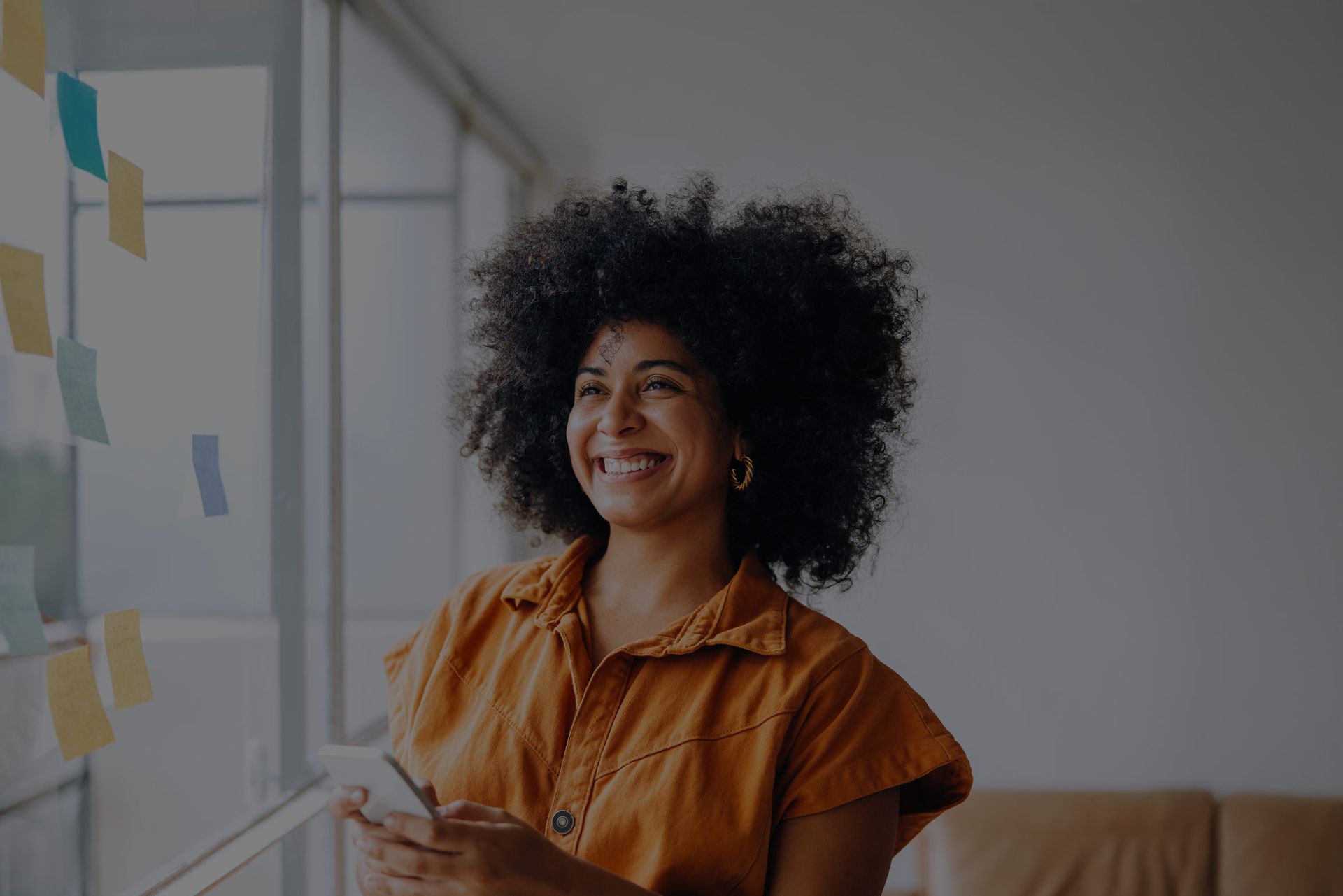 Woman with curly hair smiles while holding a phone near a window with sticky notes.