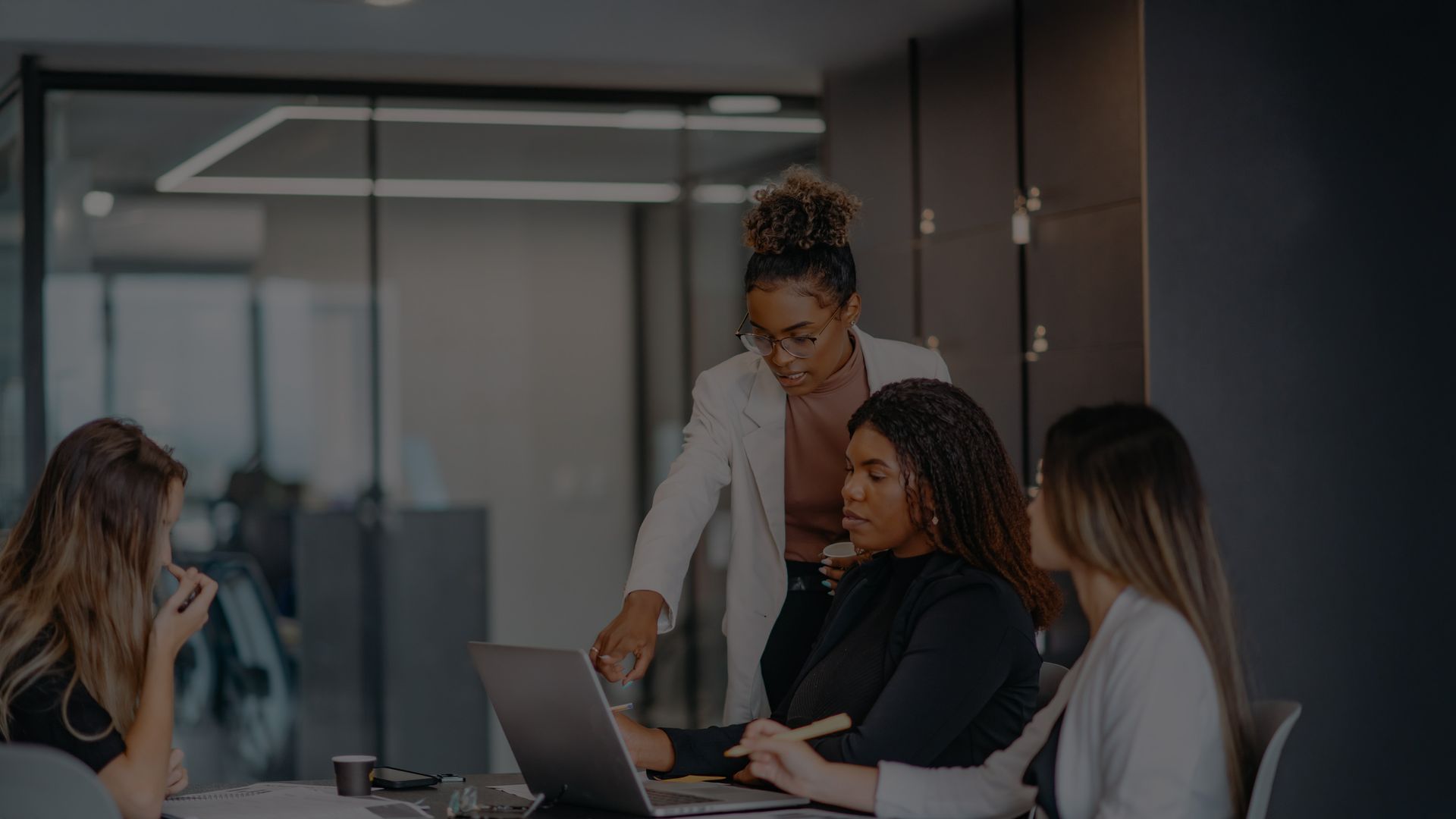 Four colleagues in a meeting, one pointing at a laptop, others looking on. Office setting.