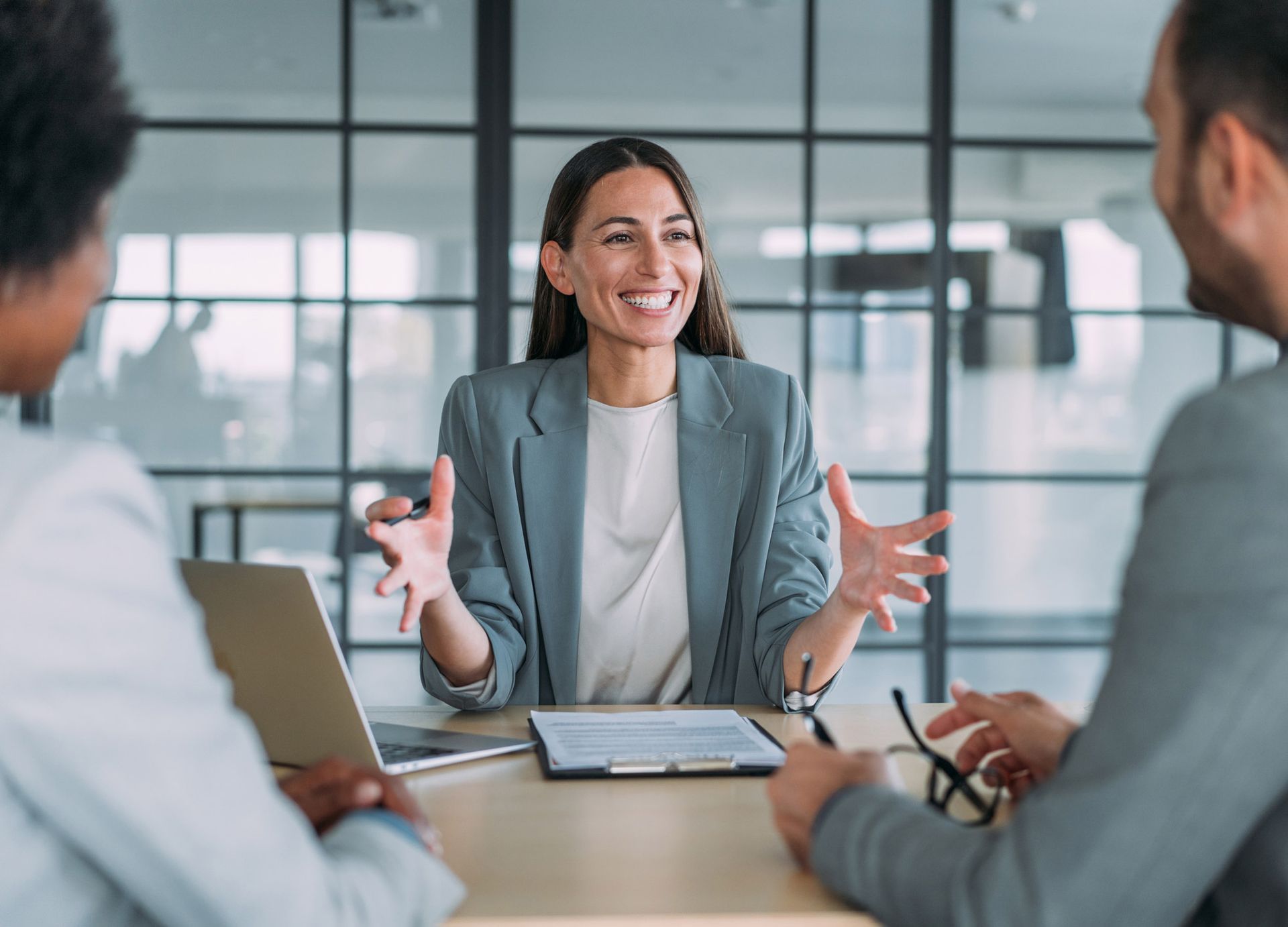Woman in gray blazer speaks animatedly, gesturing with hands, seated at a table with two colleagues in a modern office.