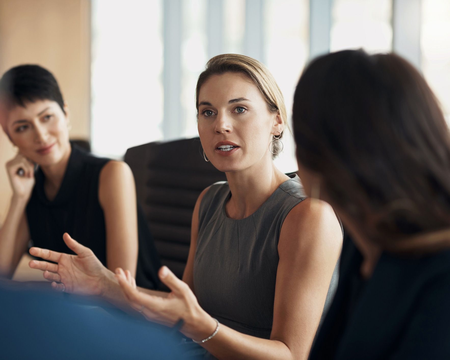 Three women in a meeting, one speaking with hands raised, two others listening attentively in office setting.