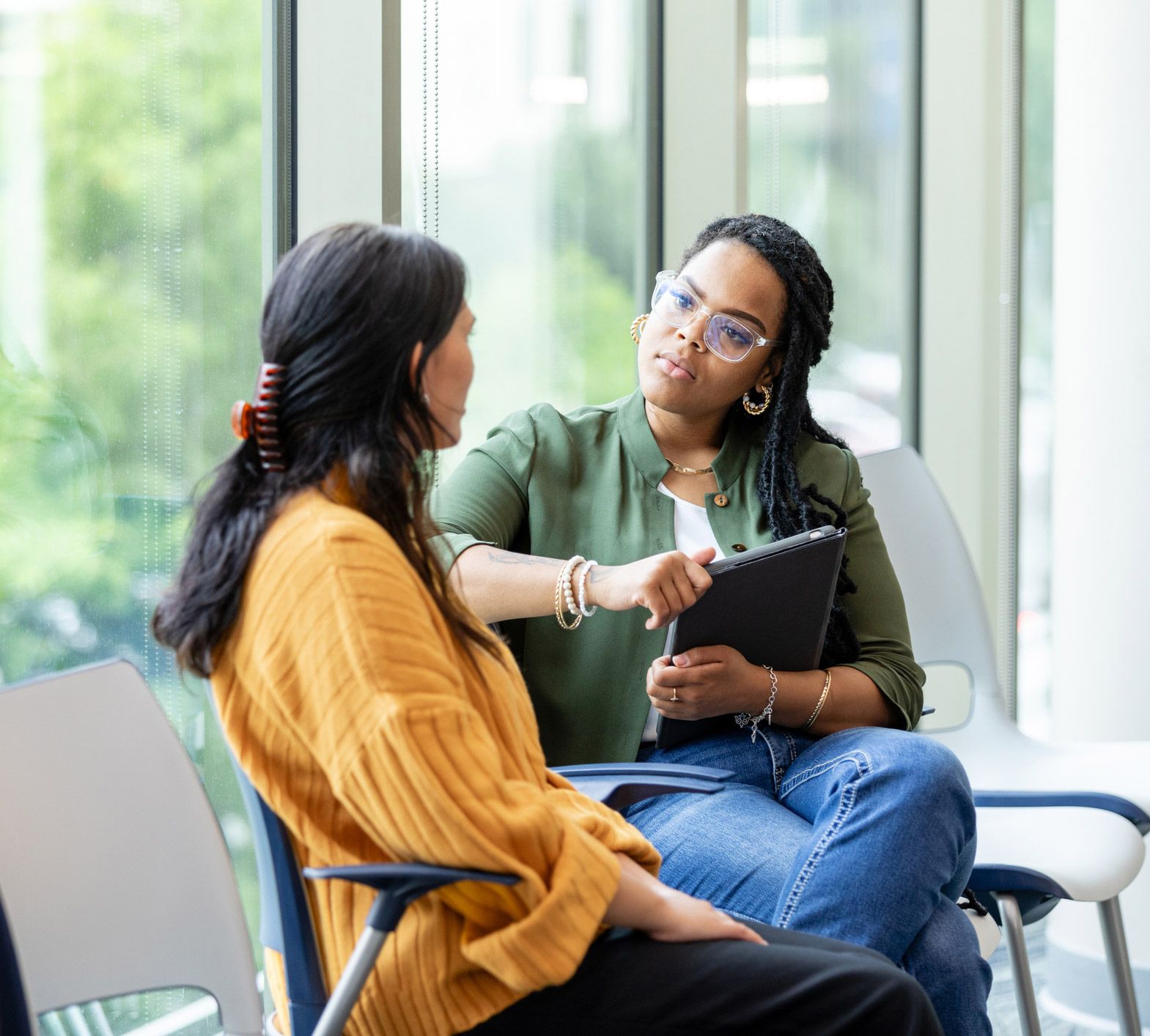 Woman in yellow top and another in green jacket talk, seated indoors. The woman in green gestures.