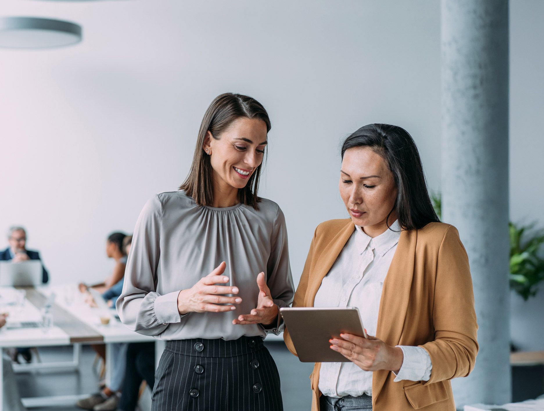 Two women in an office looking at a tablet, one pointing and smiling, in a bright, modern setting.