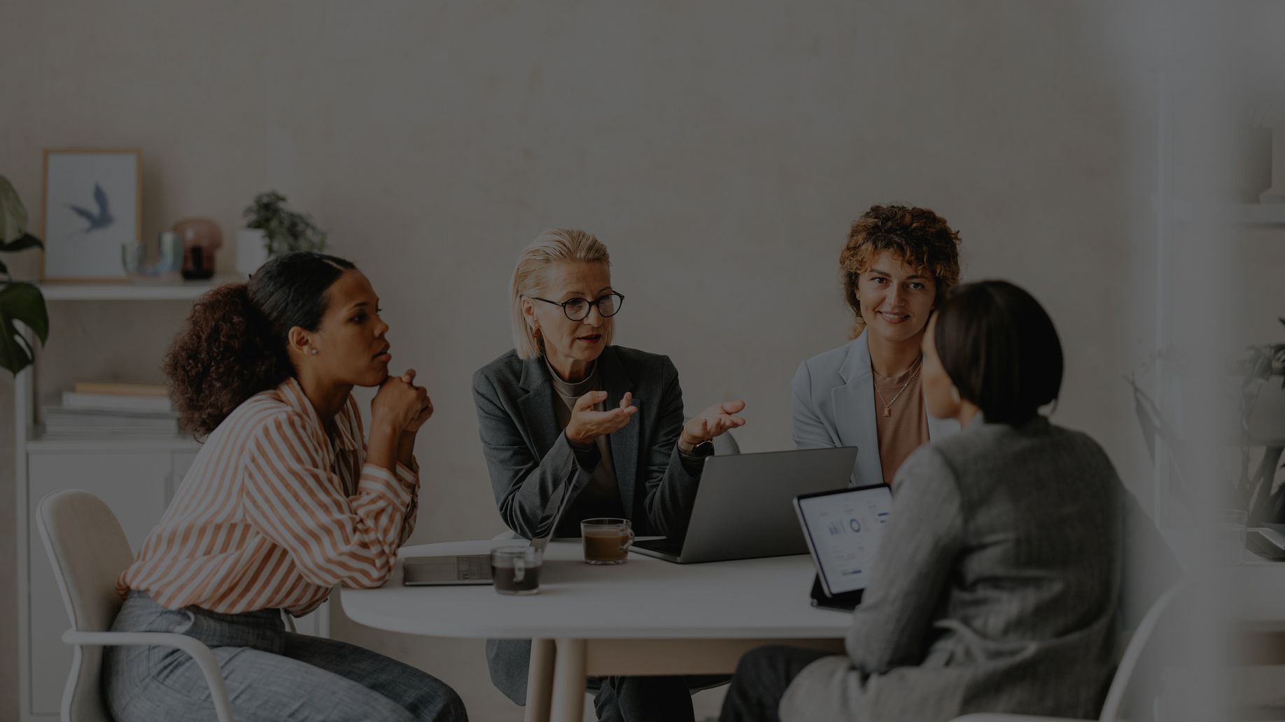 Four people in discussion around a table, one using a laptop, in a bright office.