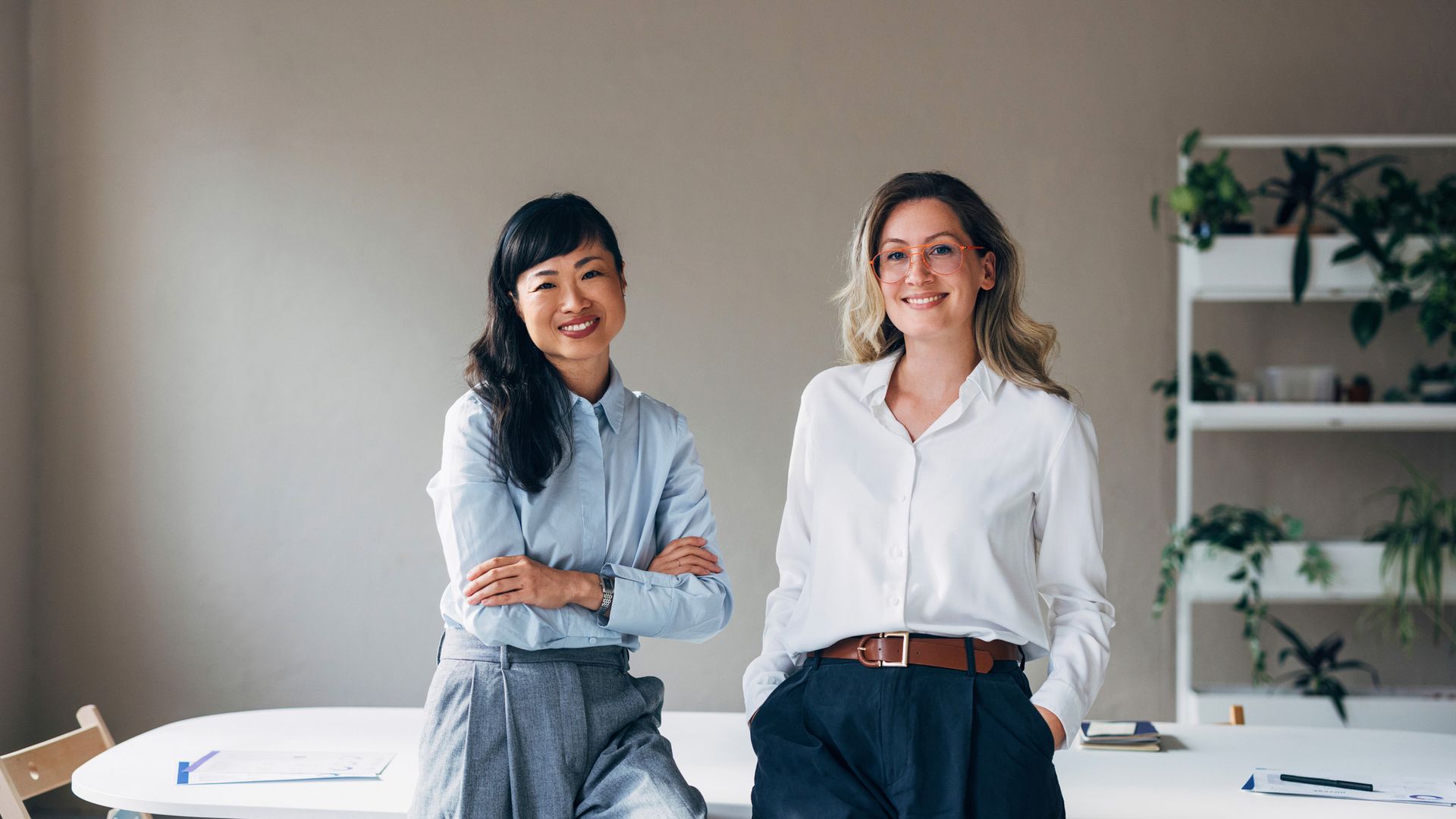 Two smiling people in office: one with arms crossed, other with hand in pocket, in front of a desk and plants.
