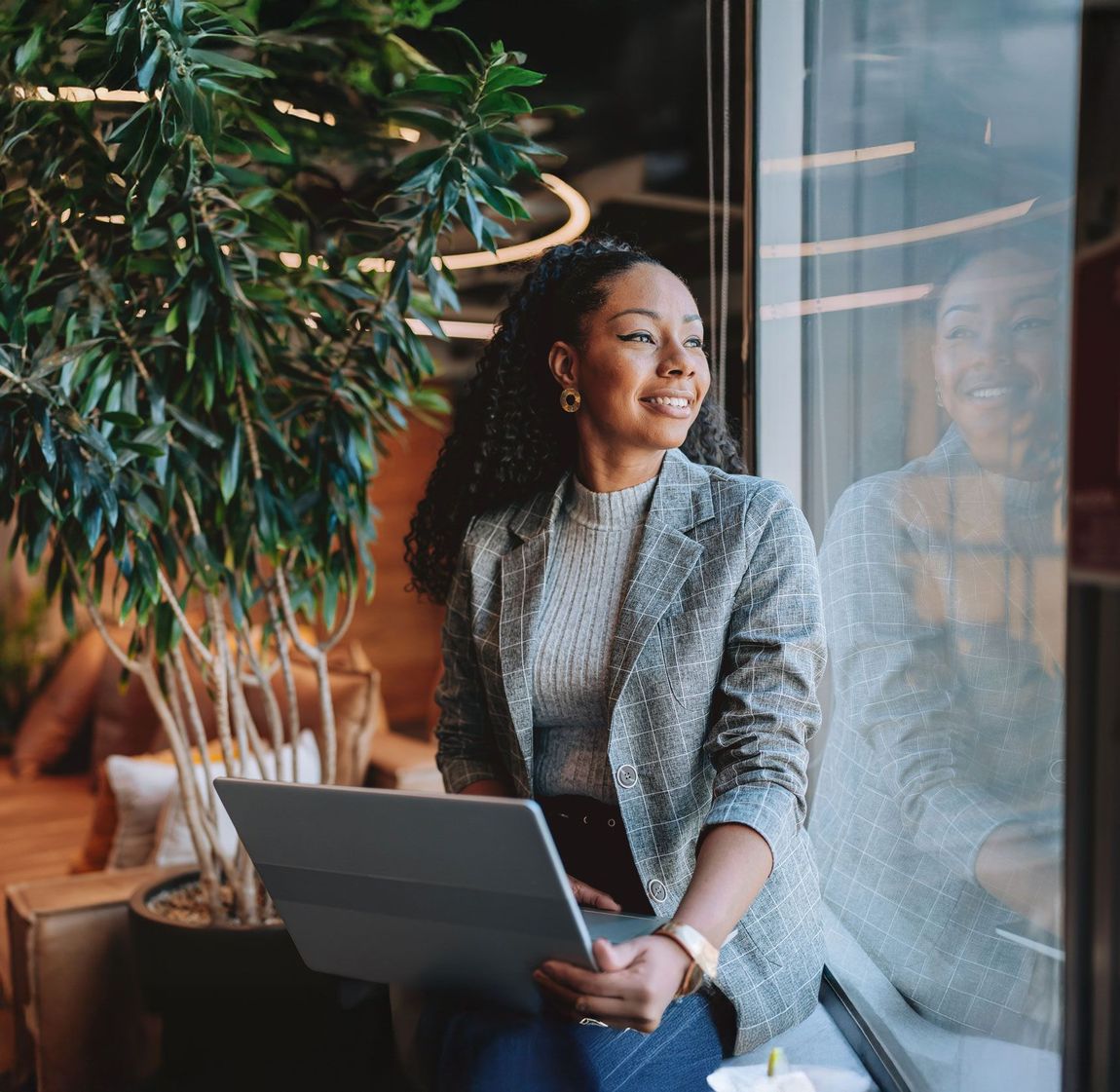 Woman in blazer smiles, looking out a window while holding a laptop, near a plant.