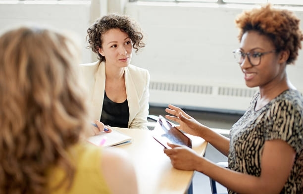 Three women at a table; one speaks, holding a tablet, the others listen. Light-filled room.