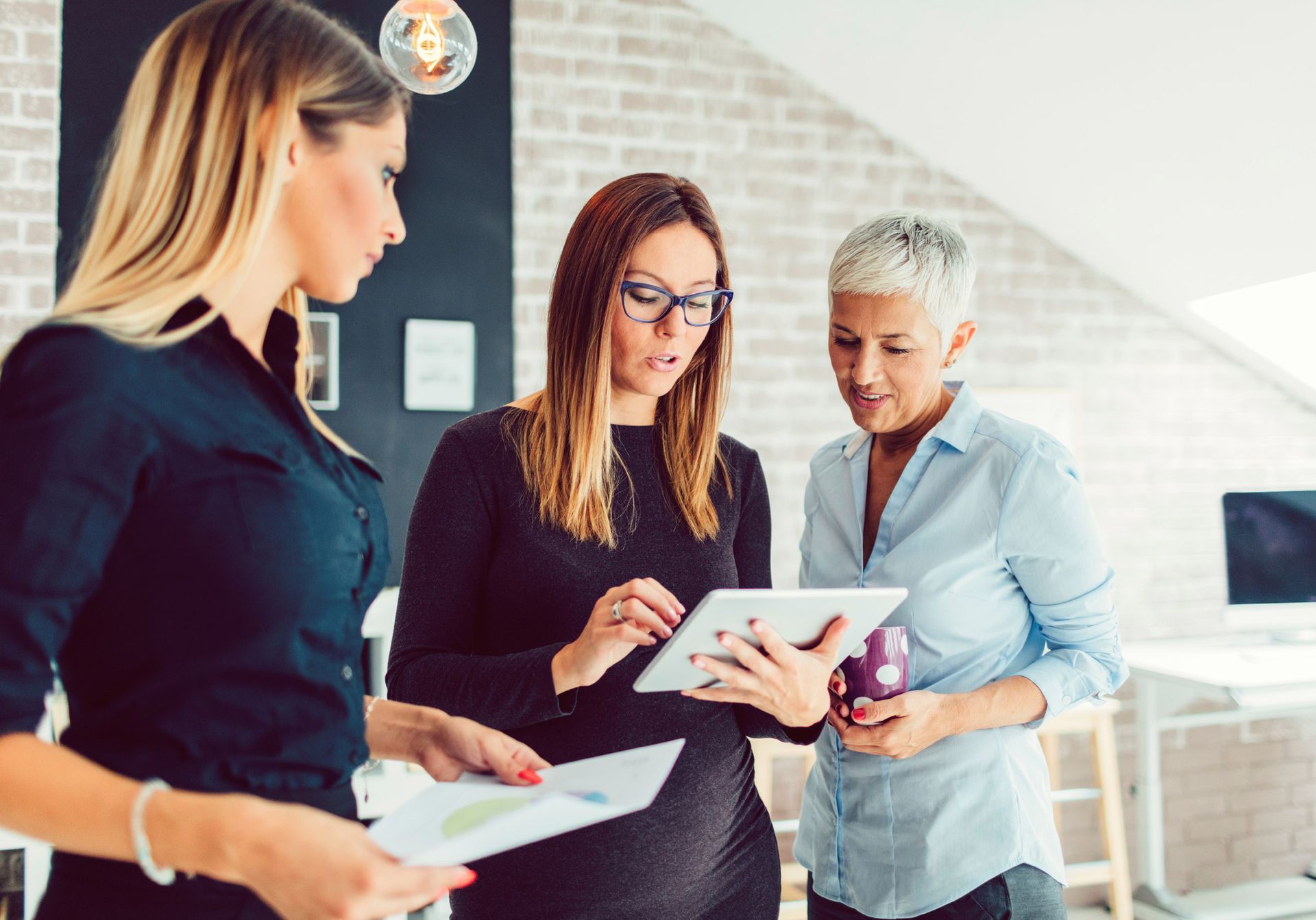 Three women looking at a tablet and documents in an office setting.