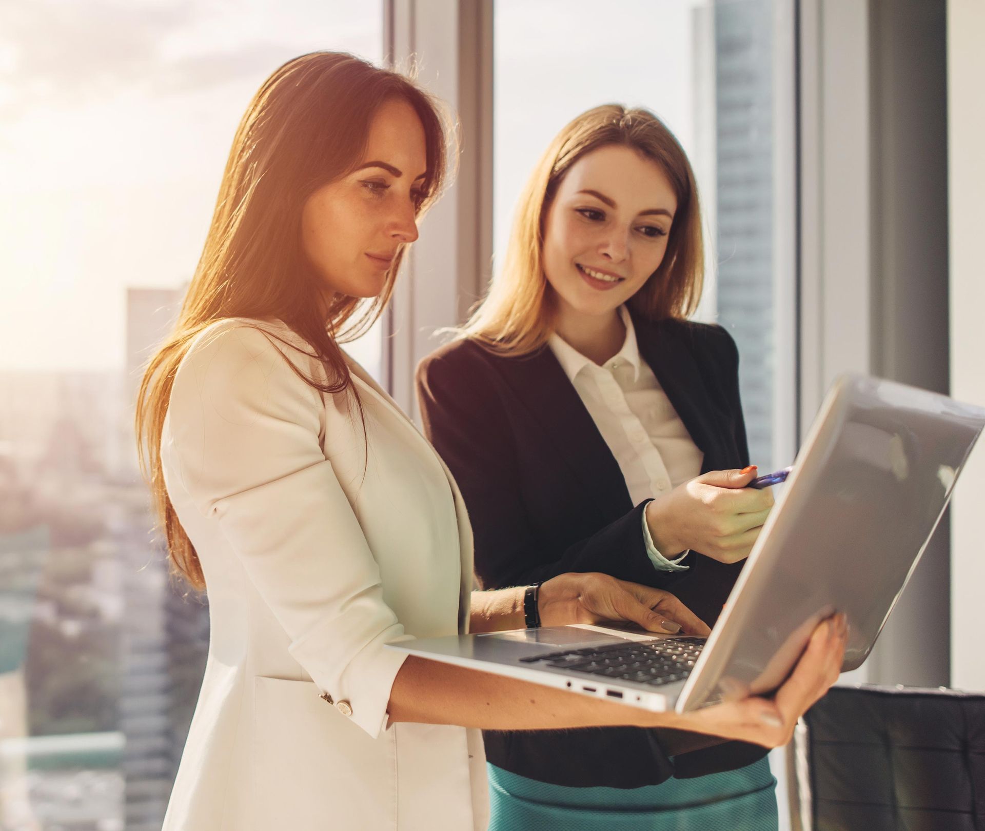 Two women in business attire looking at a laptop in a bright office, one pointing at the screen.