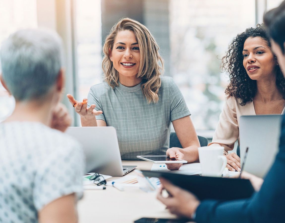 Business meeting with smiling woman speaking, surrounded by colleagues and laptops.
