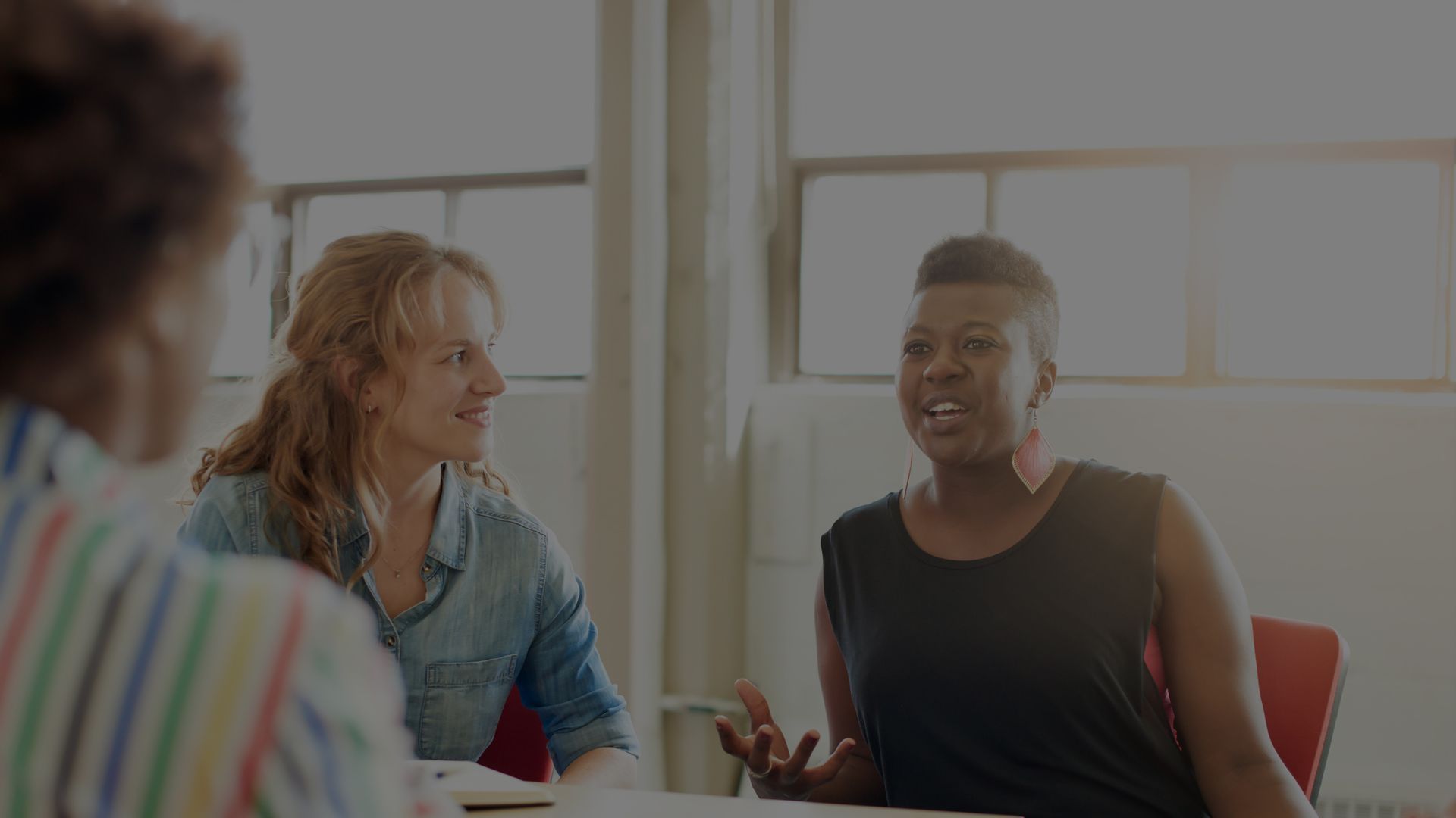 Three people in a meeting, one woman speaking with hand gestures, others listening attentively. Bright room, natural light.