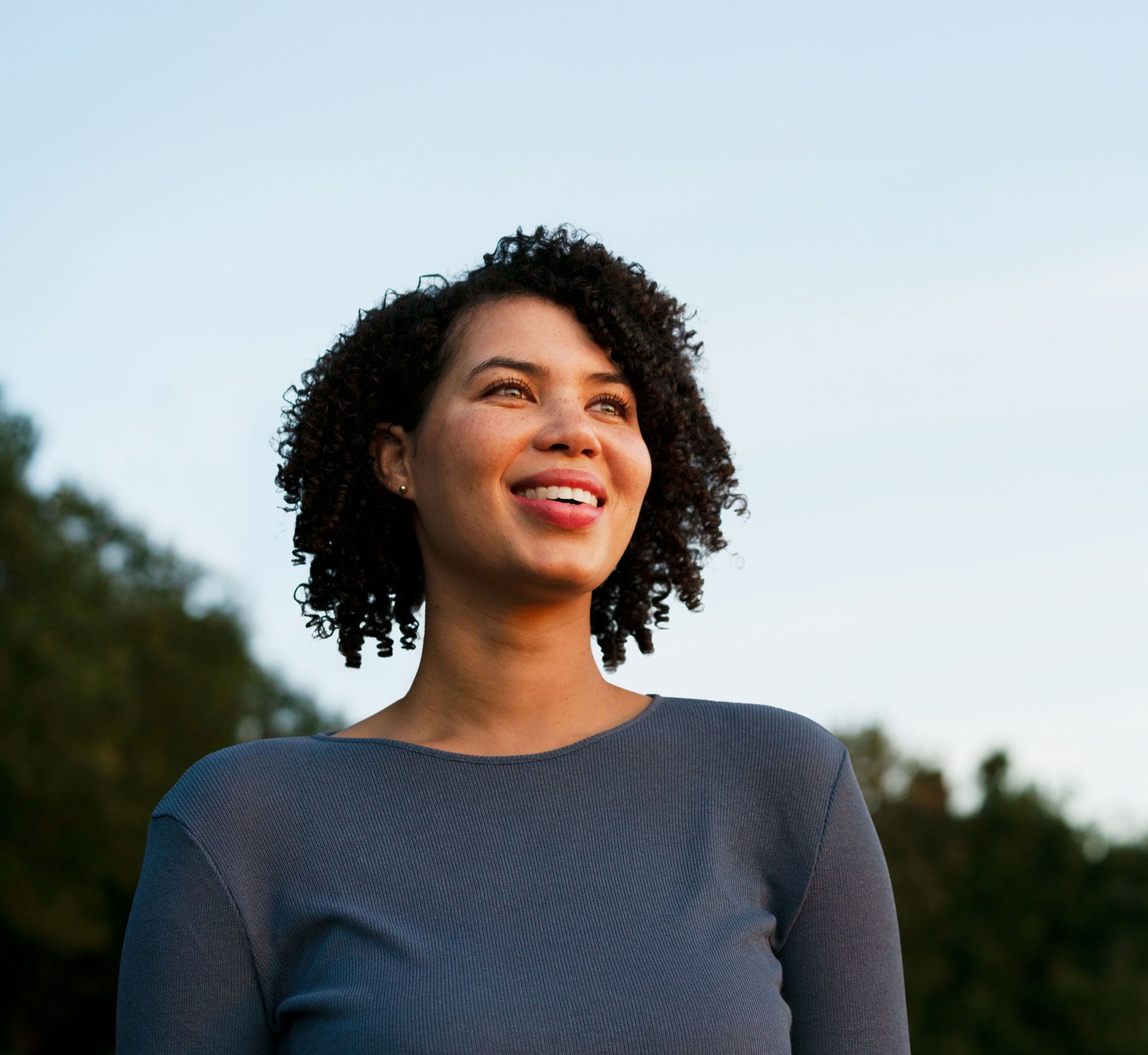 Woman with curly hair smiles, looking upwards, outdoors with a blue sky background.