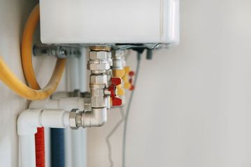 Close-up of a white residential water heater with metal piping, yellow hoses, and red and yellow valve handles.