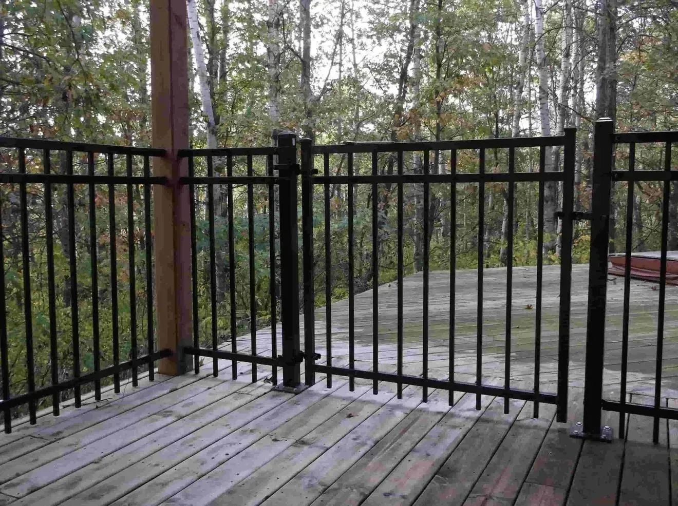 Black metal fence with gate on a wooden deck, forest in background.